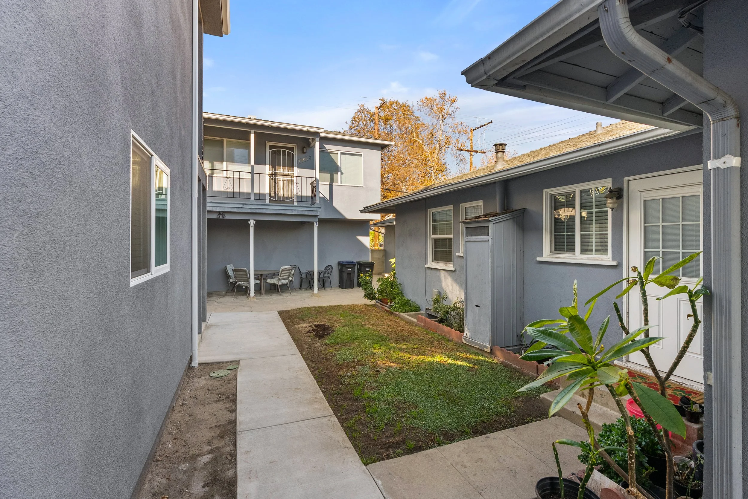 A small outdoor area with a concrete pathway, a patch of dirt, and a grassy section, bounded by gray house walls with windows, a backyard patio with a table, chairs, and trash bins, and a building with a balcony in the background.