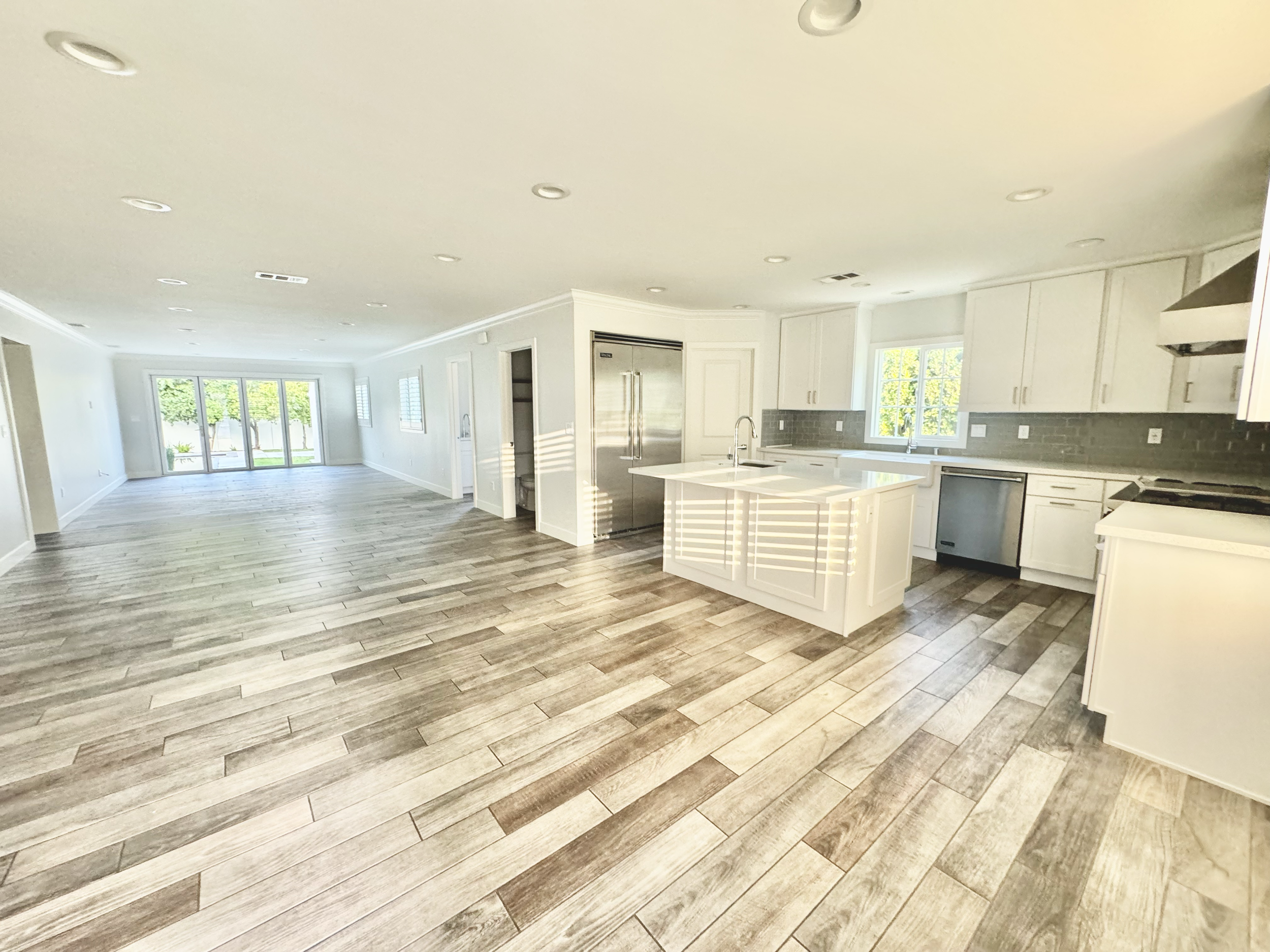 Open floor plan living space with kitchen, white cabinets, gray backsplash, large windows, hardwood floors, sliding glass doors leading to backyard.