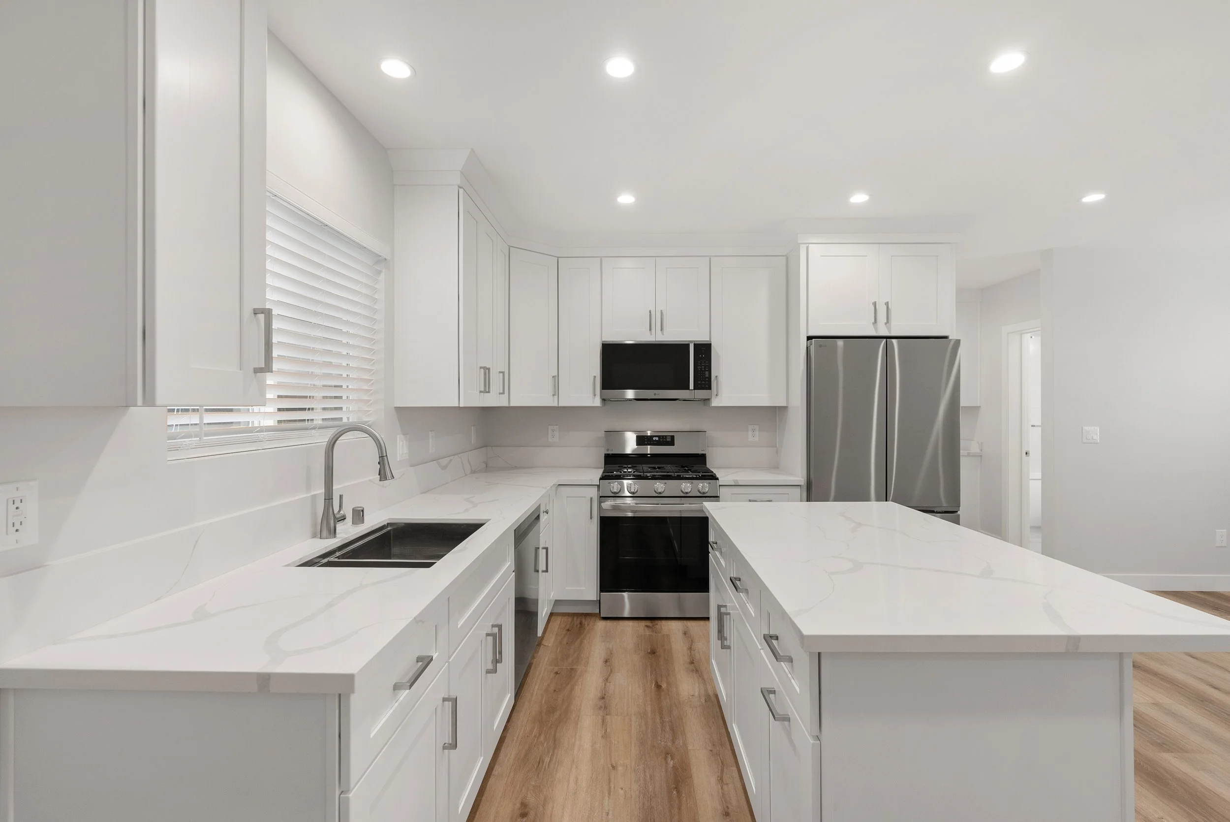 Modern white kitchen with marble countertops, stainless steel appliances, hardwood flooring, and recessed ceiling lights.