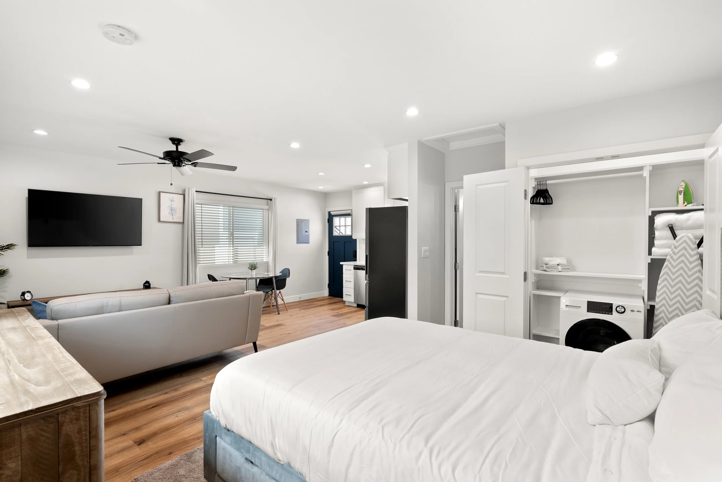 Bright, modern studio apartment featuring a bed, a washer and dryer in a closet, a gray sofa, a flatscreen TV, a small dining table with chairs, hardwood floors, white walls, and ceiling lights.