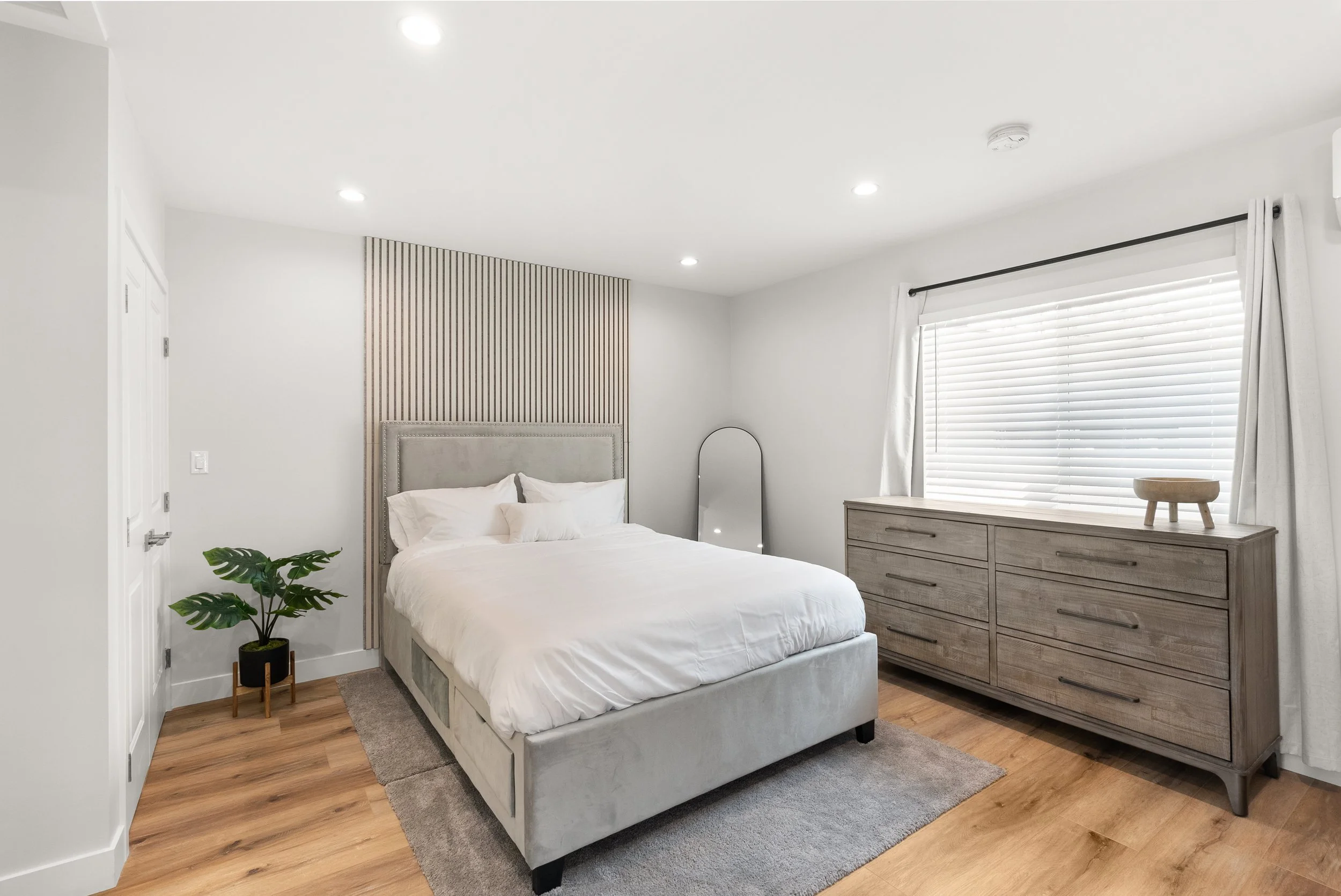Minimalist bedroom with a gray bed, wooden dresser, green plant, mirror, and window with white blinds.