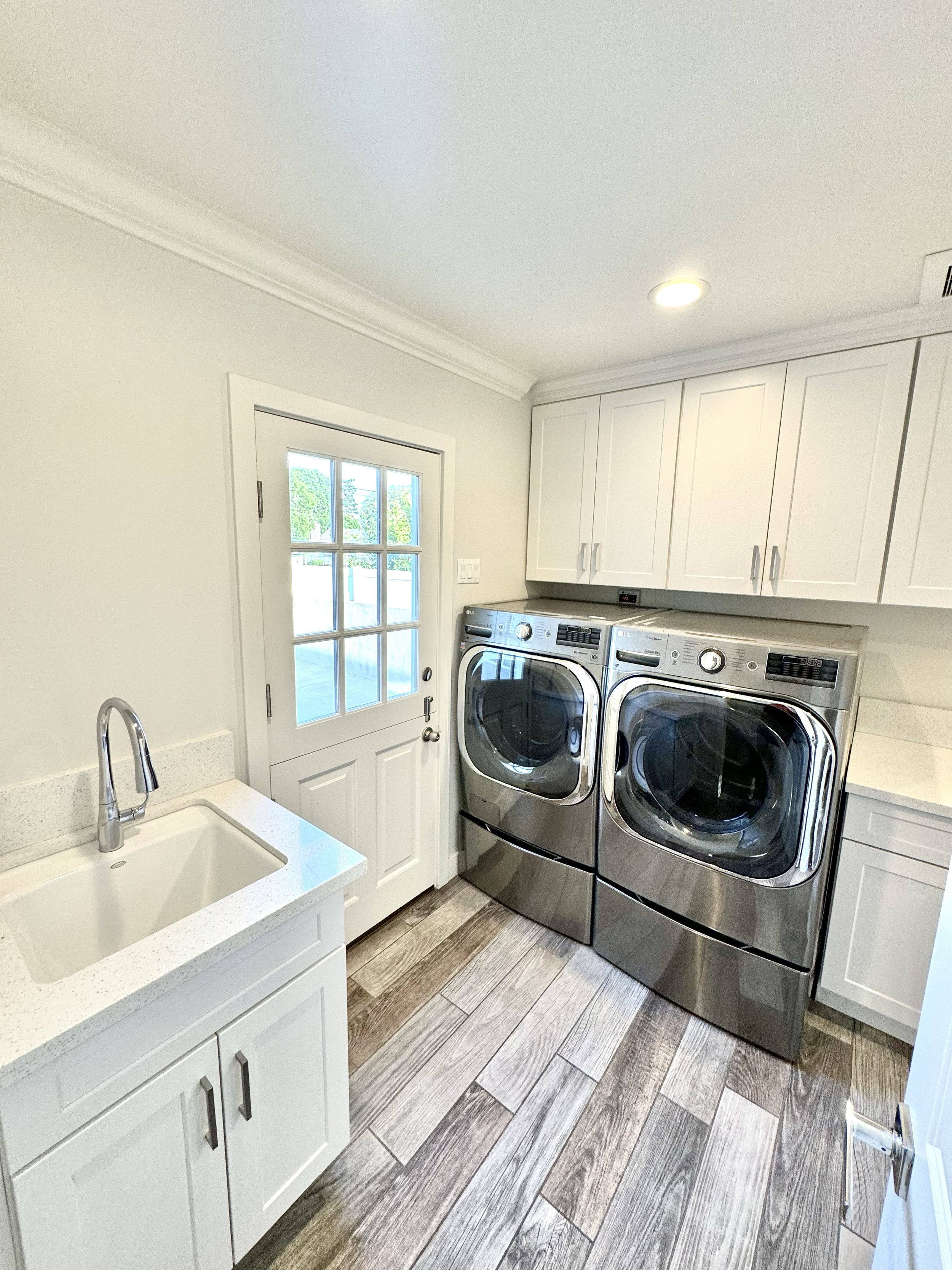 Laundry room with front-loading washer and dryer, white cabinets, a small utility sink, and a door with glass panes leading outside.