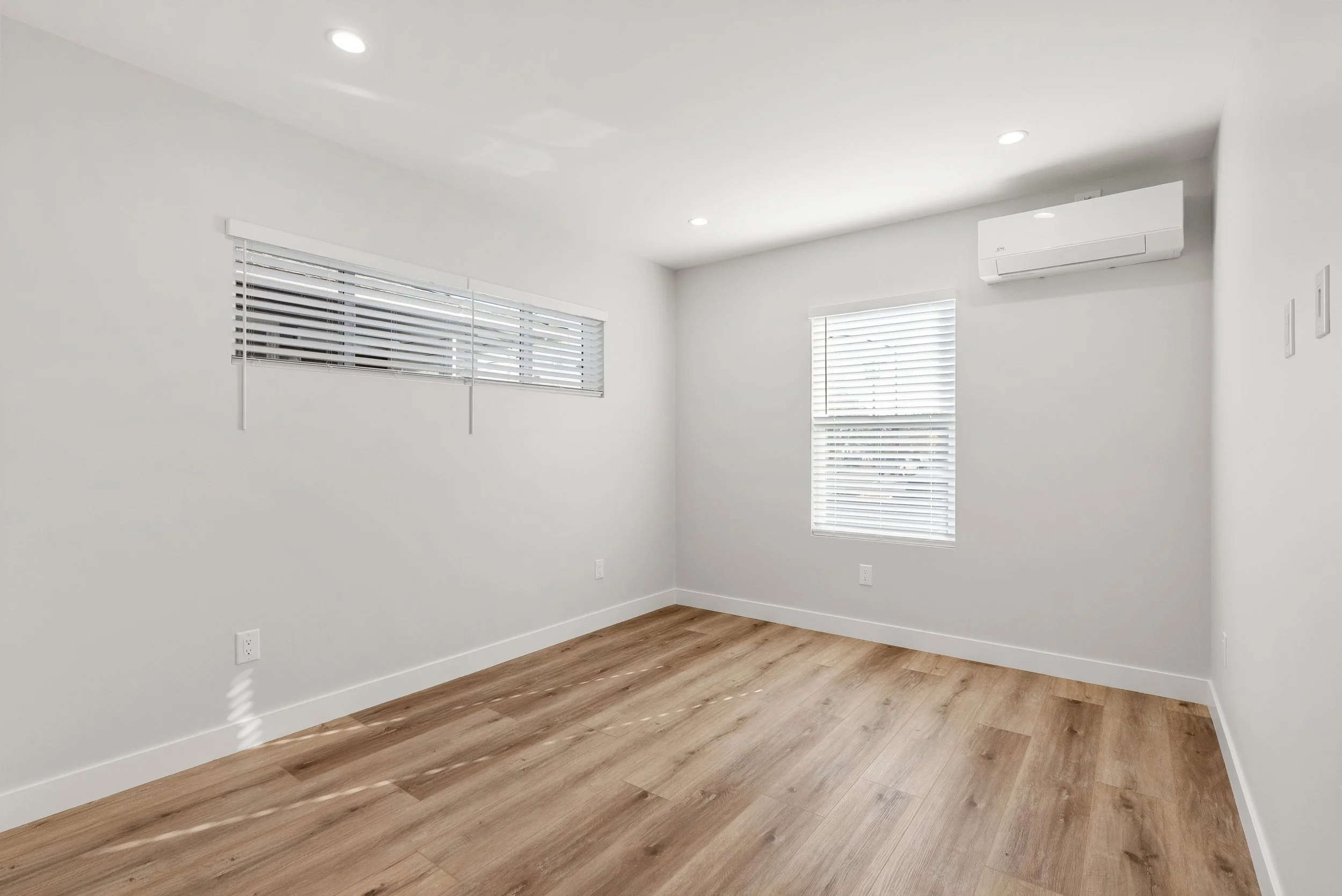 Empty room with white walls, hardwood flooring, three windows with white blinds, and an air conditioning unit on the wall.