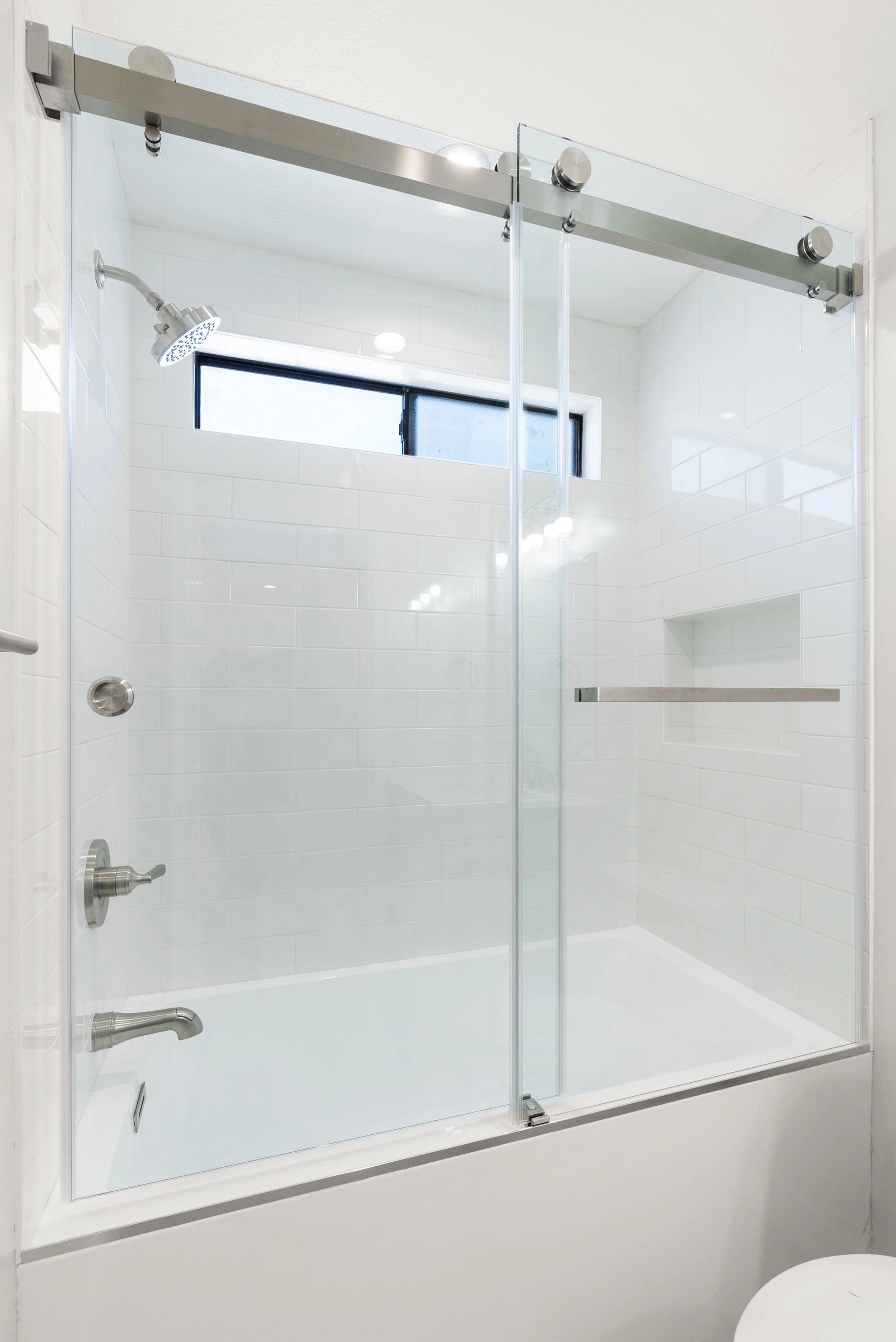 A modern glass shower enclosure with a sliding door, stainless steel fixtures, white subway tile walls, a small built-in shelf, and a frosted window