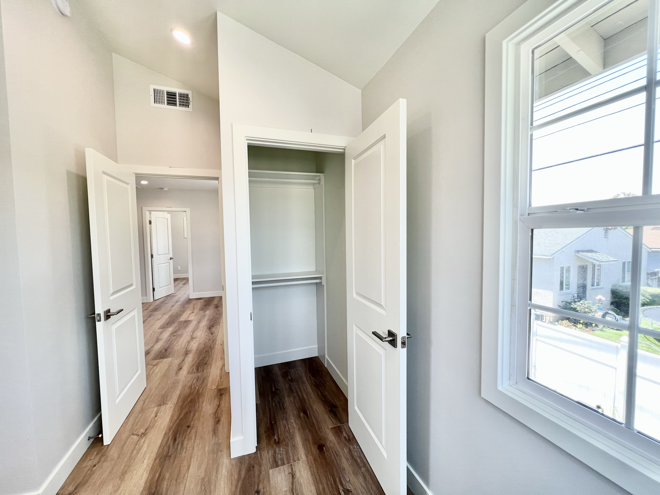 Empty small closet with open door, white walls, hardwood flooring, and window showing neighborhood houses.