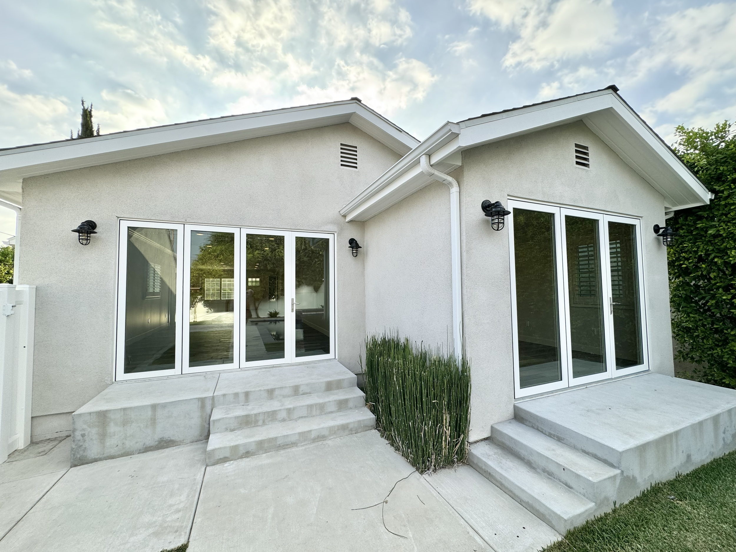 Rear view of a modern house with two sets of glass sliding doors and outdoor lighting fixtures, concrete steps, and a small patch of grass, under a partly cloudy sky.