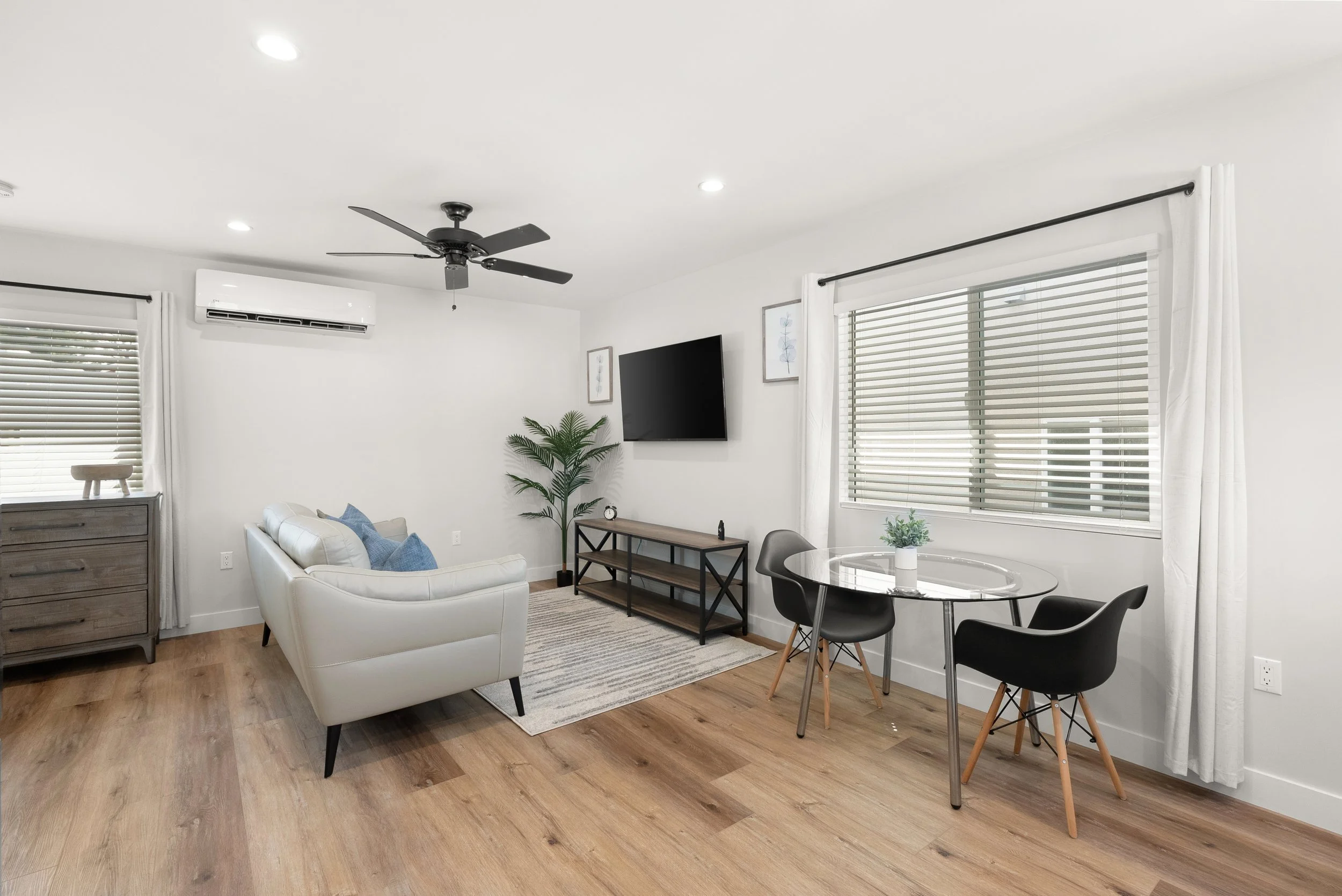 Living room with white walls, hardwood floors, a white sofa with blue pillows, a small gray dresser, a potted plant, a wall-mounted TV, a black ceiling fan, two framed art pieces, a glass dining table with black chairs, and large windows with white c