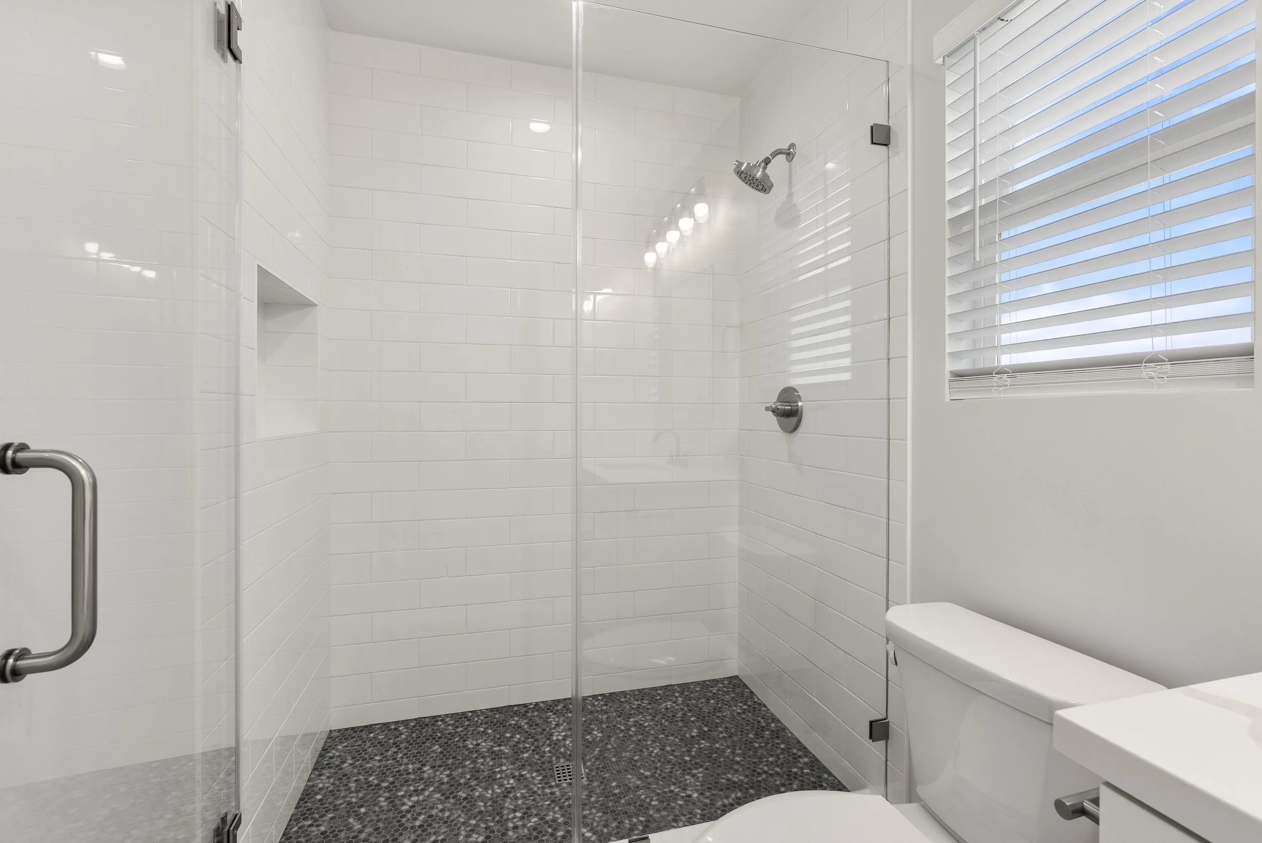 Modern bathroom with a walk-in shower, white subway tile walls, a window with white blinds, and a white toilet.