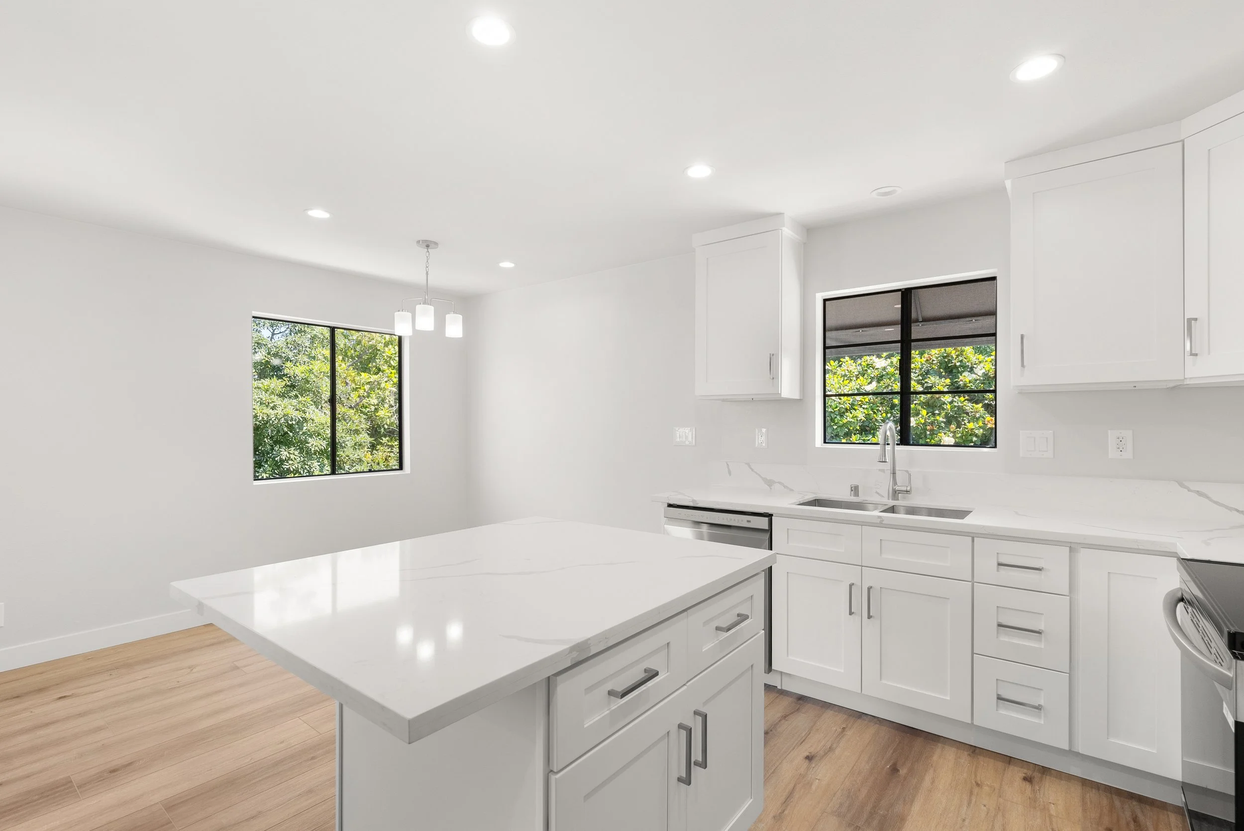 Empty white kitchen with island, white cabinets, stainless steel sink, window showing green foliage outside, hardwood floor, and overhead lighting.