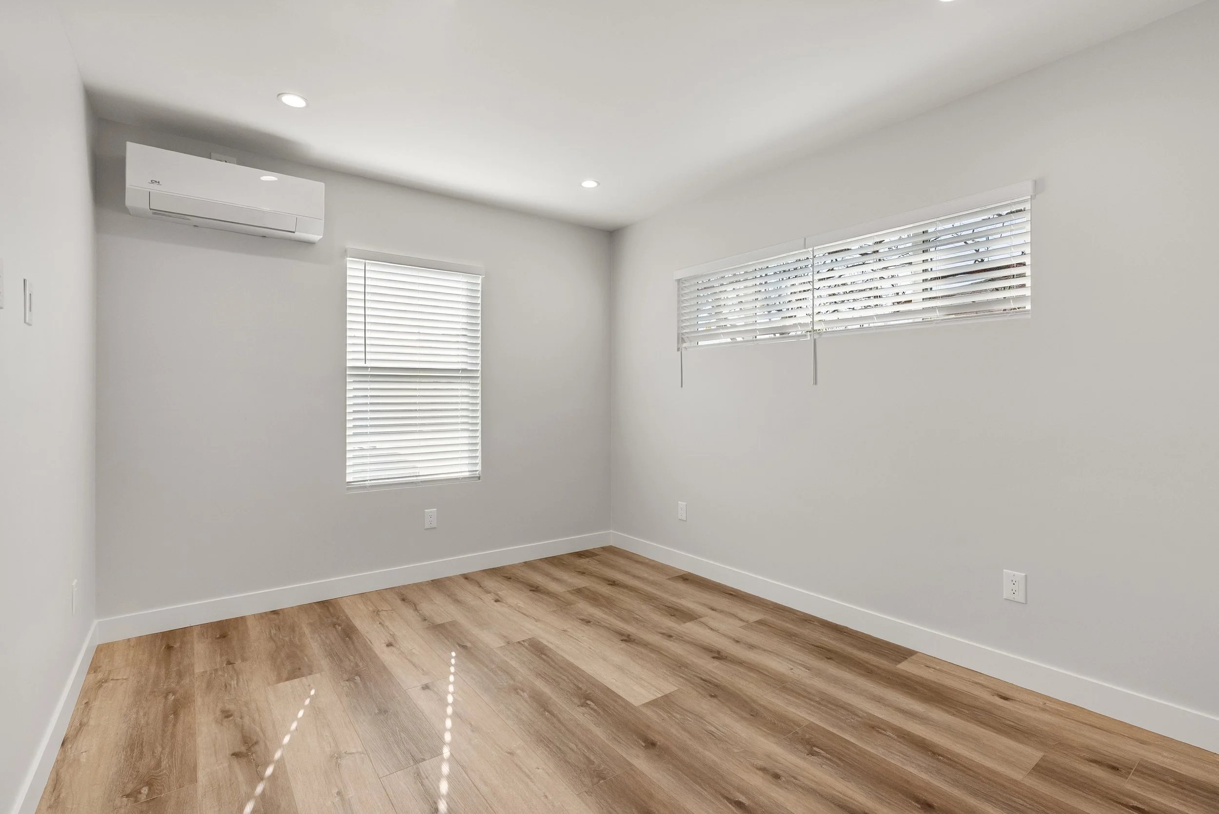 Empty room with white walls, wood flooring, two windows with blinds, and an air conditioner on the wall.
