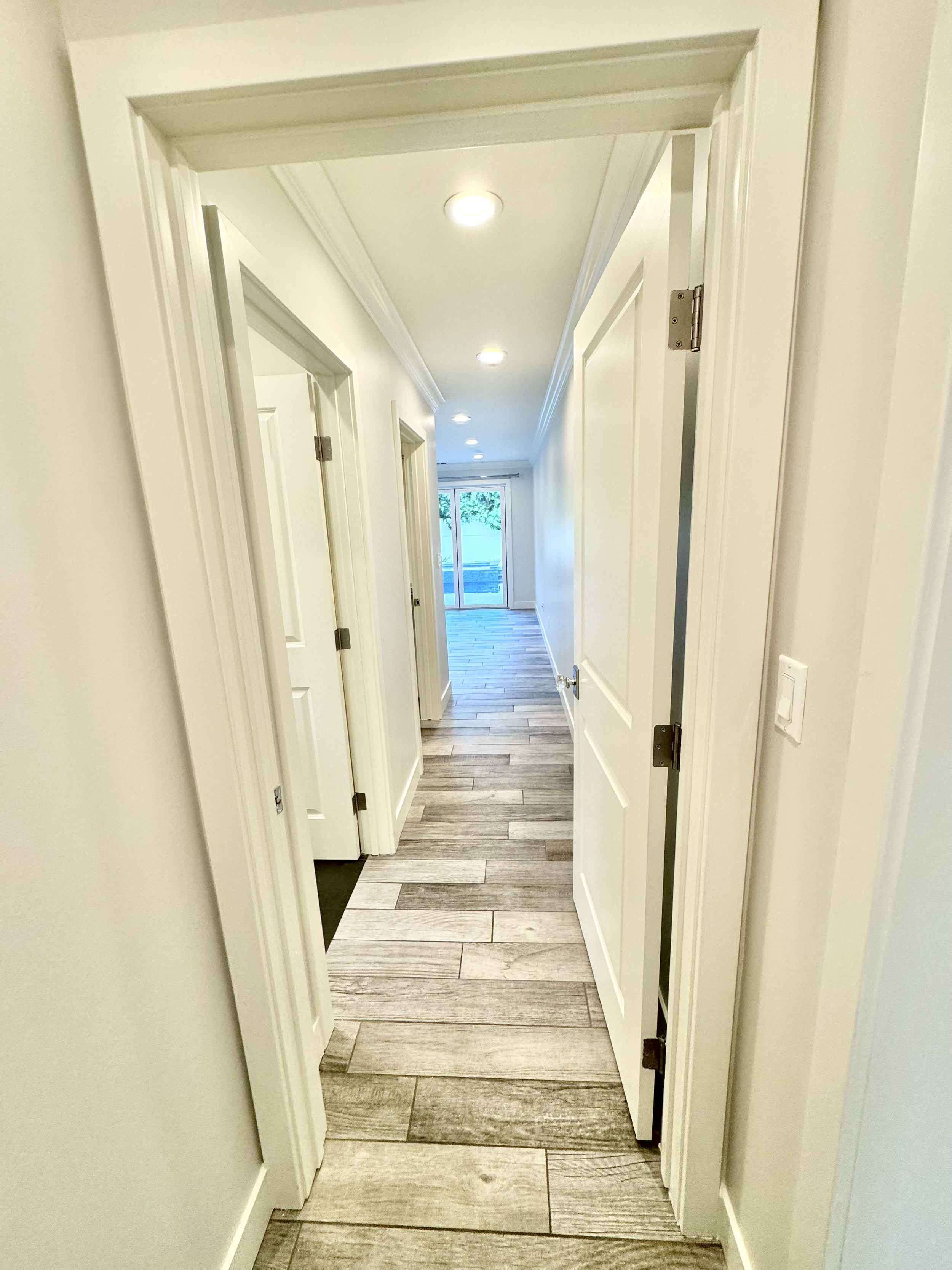 Empty hallway with wooden flooring, white walls, and multiple closed doors, leading to a sliding glass door at the end.