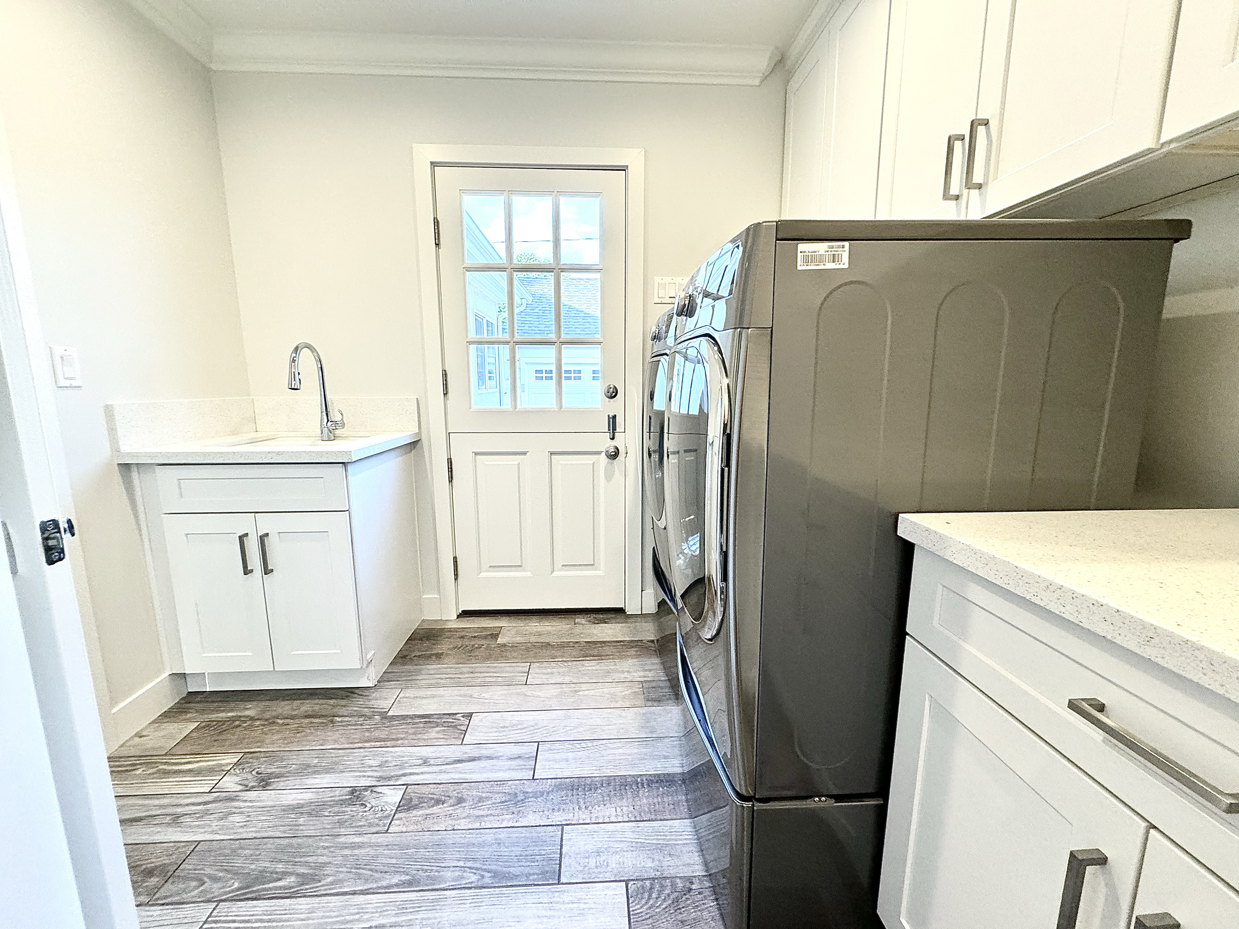 Laundry room with white cabinets, a wash basin, a door with window panes showing an outdoor view, a sink with a faucet, laundry appliances, and wooden plank flooring.