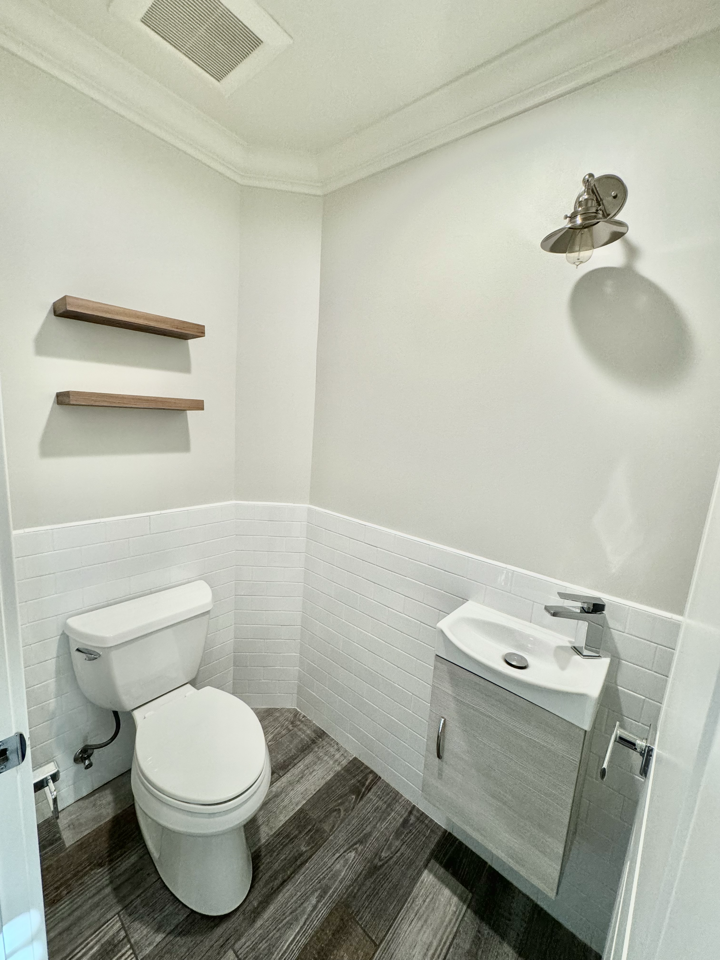 A small bathroom with a white toilet, a wall-mounted small sink with a cabinet, two wooden shelves, a wall light, and wood-style flooring.