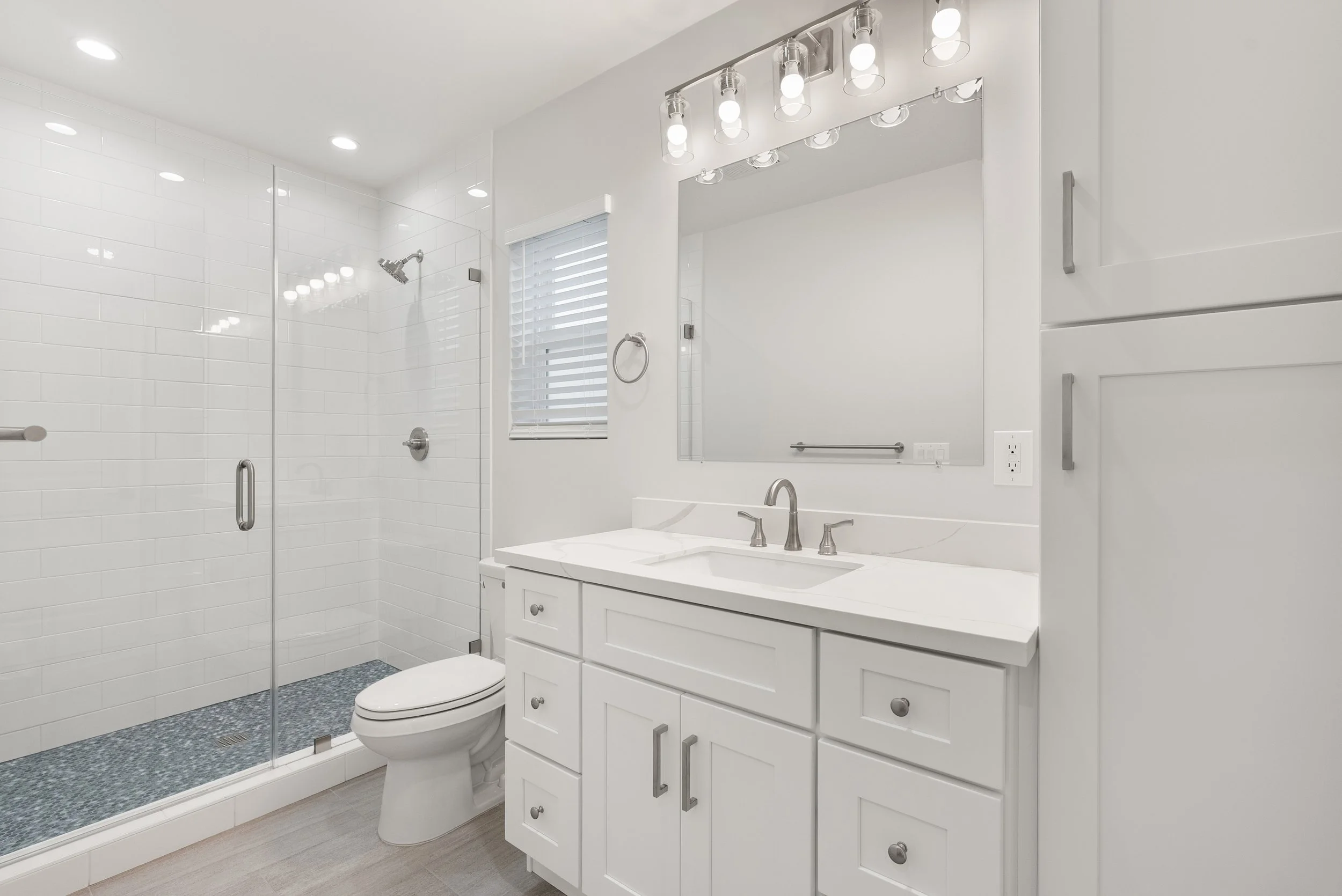 Modern white bathroom with a glass-enclosed shower, white vanity, large mirror, and a window with blinds.