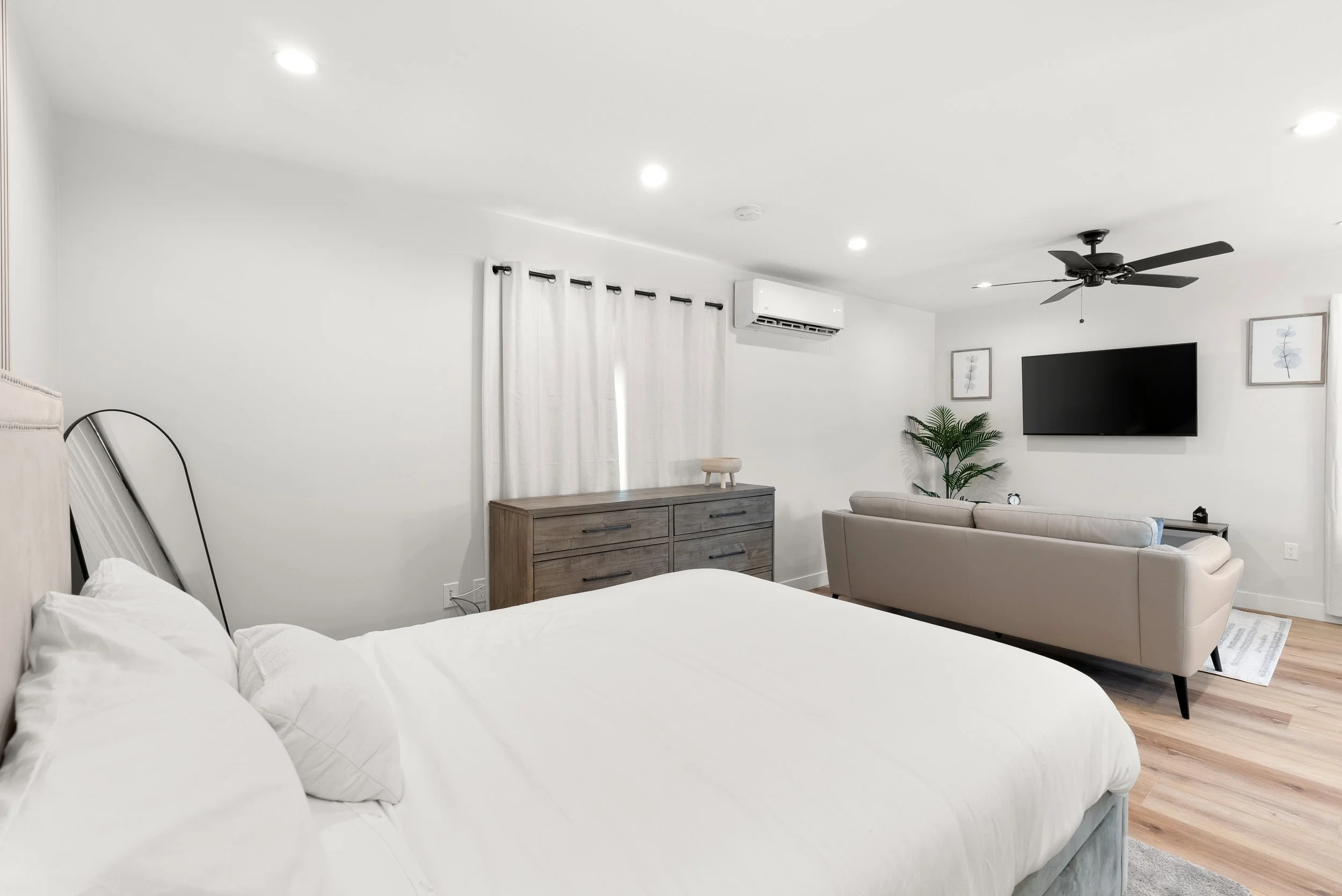 Minimalist bedroom with a large bed, dresser, beige sofa, wall-mounted TV, ceiling fan, and wall art, with natural light and white curtains.