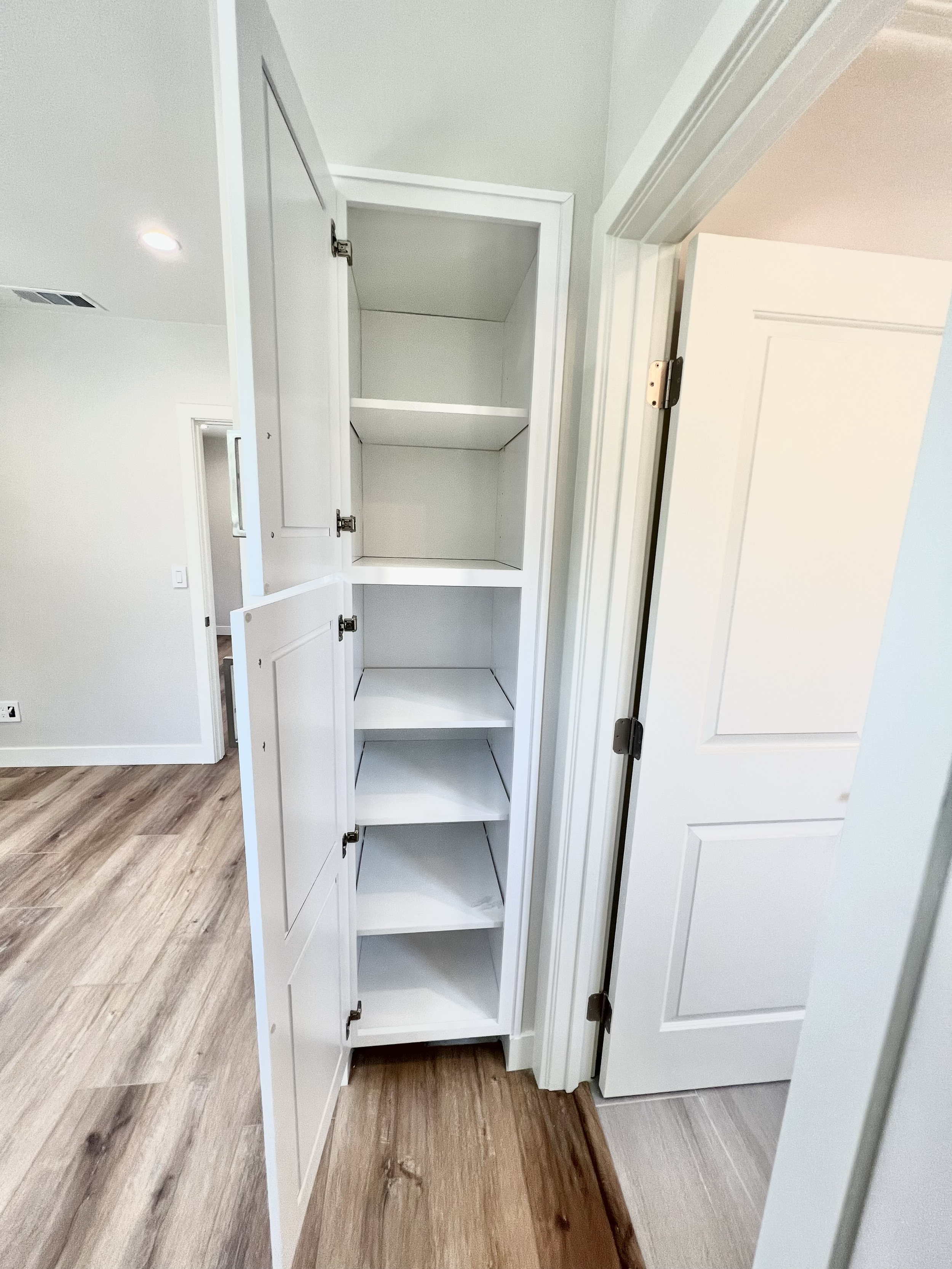 Open white built-in pantry cabinet with four empty shelves in a bright room with light-colored walls and wood laminate flooring.