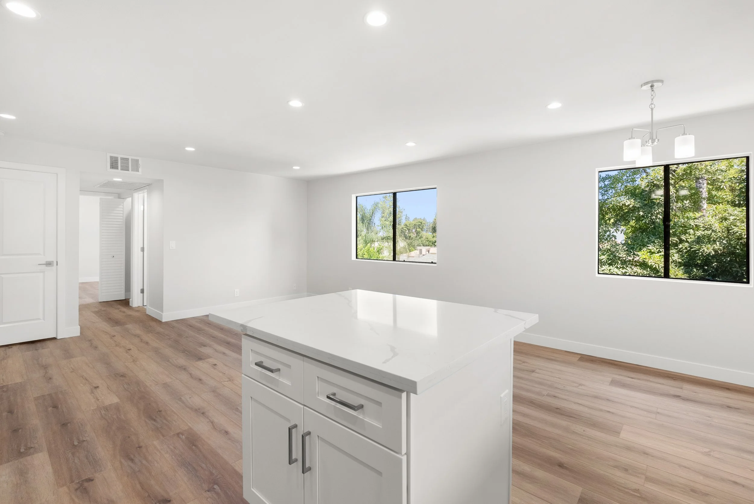 Empty modern kitchen with white walls, ceiling, island, wood flooring, windows showing green trees, and ceiling lights.