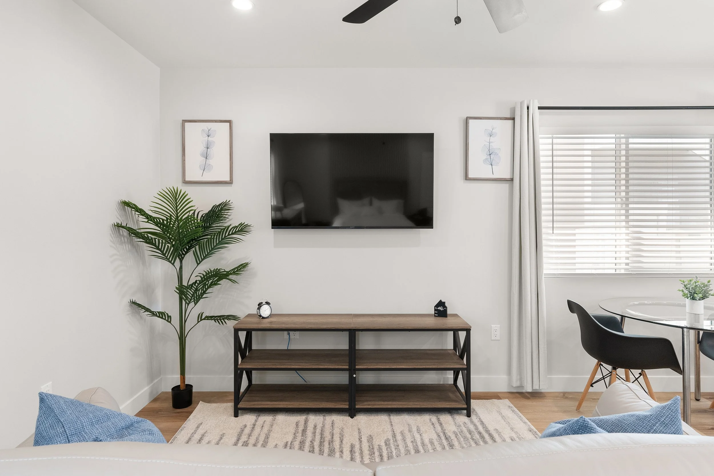 Living room with a wall-mounted TV, wooden console table, potted plant, framed wall art, window with blinds and curtains, dining table with black chairs, and a white sofa with blue pillows.