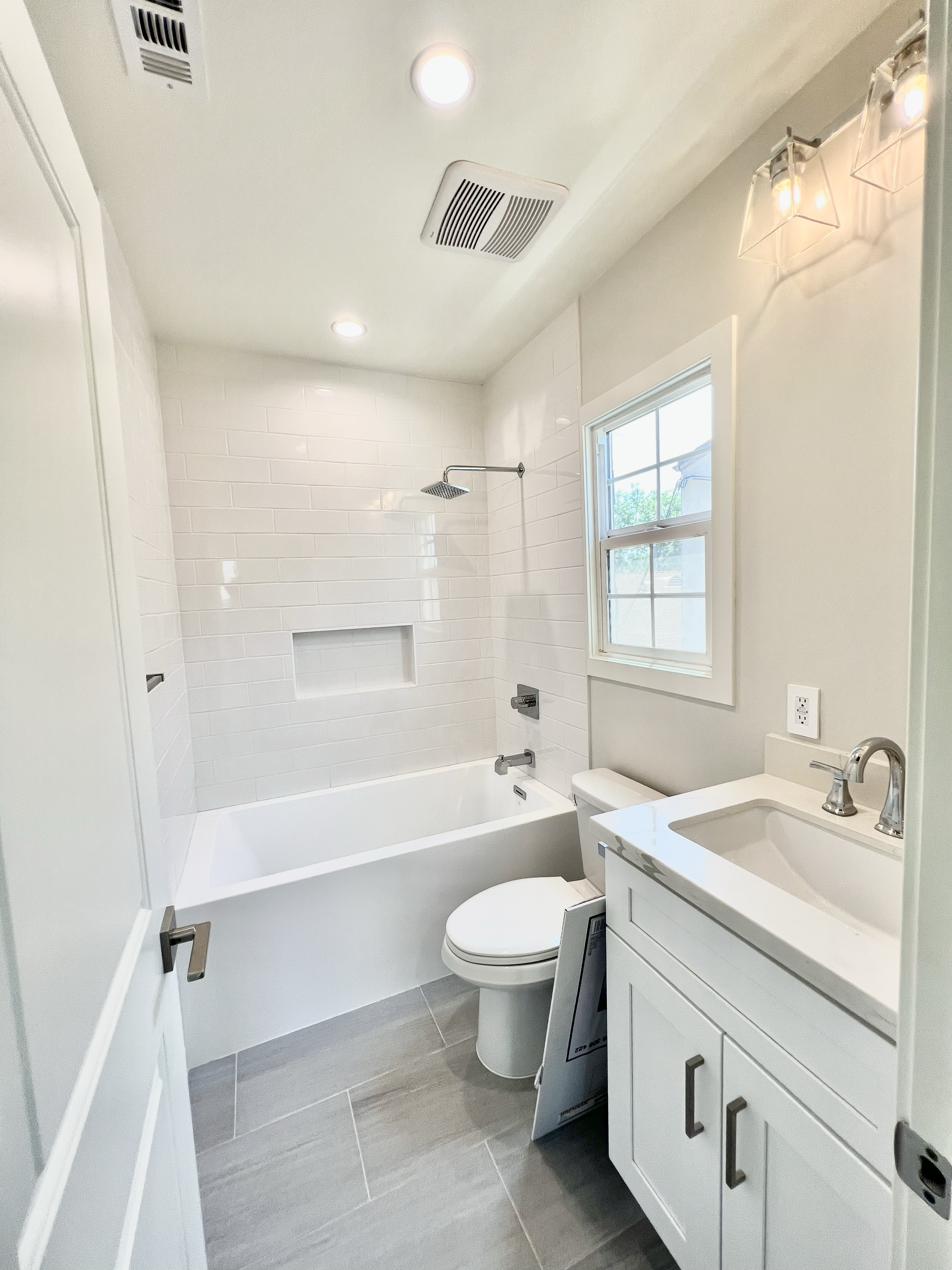 A modern bathroom with a bathtub, toilet, and small vanity with a sink. White tile surrounds the bathtub area, and a window provides natural light. Recessed and wall-mounted light fixtures illuminate the space, and there is an ventilation fan on the 