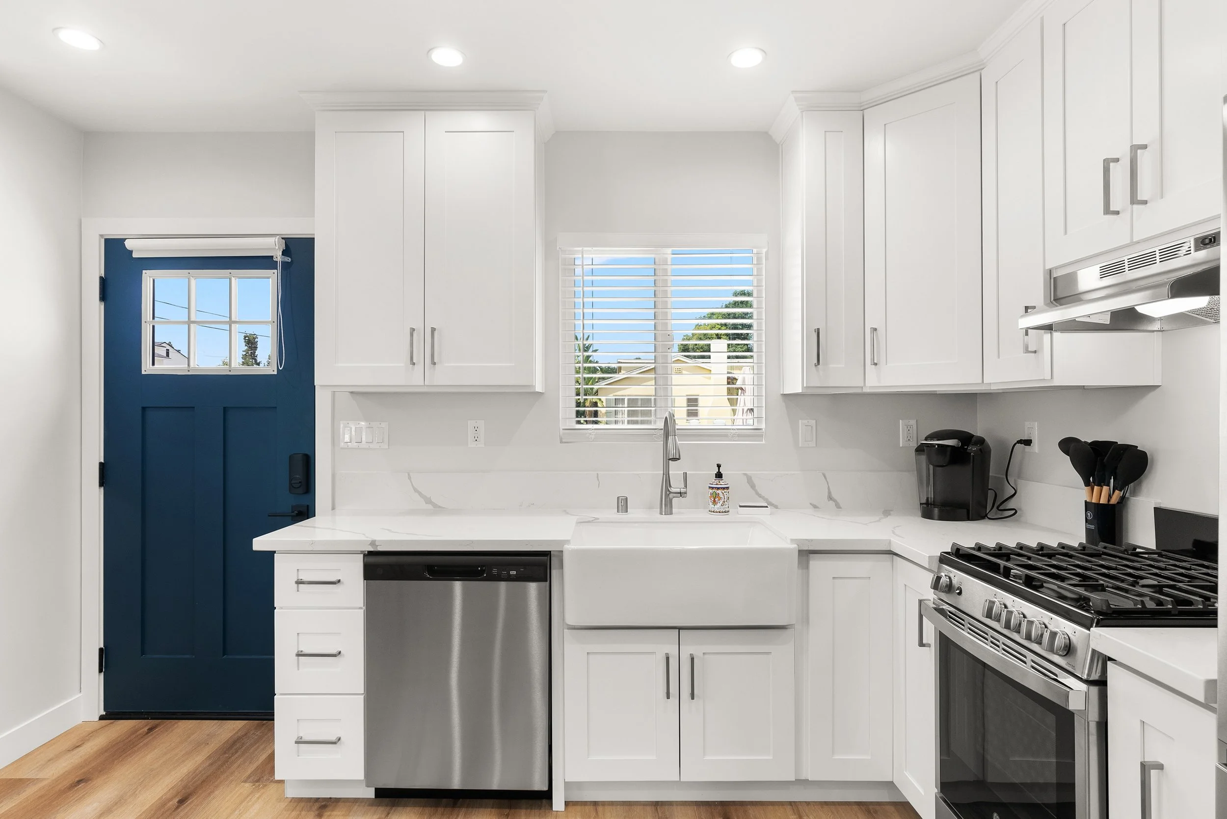 Modern white kitchen with a window above the farmhouse sink showing outdoor houses and trees, a blue door to the outside, stainless steel appliances including a dishwasher and a stove, and kitchen utensils on the counter.