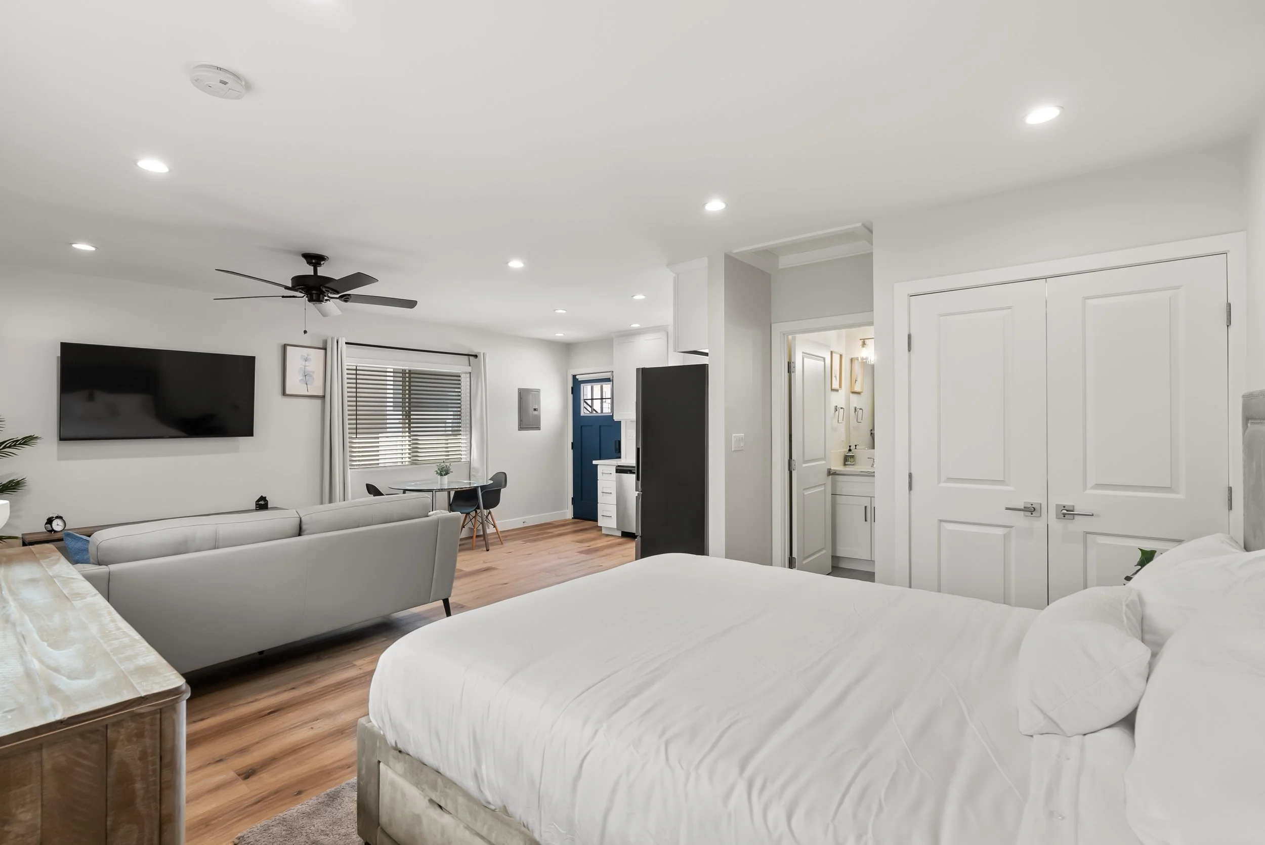 Modern living room with white walls, hardwood flooring, a mounted flat-screen TV, light gray sofa, bed with white linens, a ceiling fan, and a small dining area near a blue door.