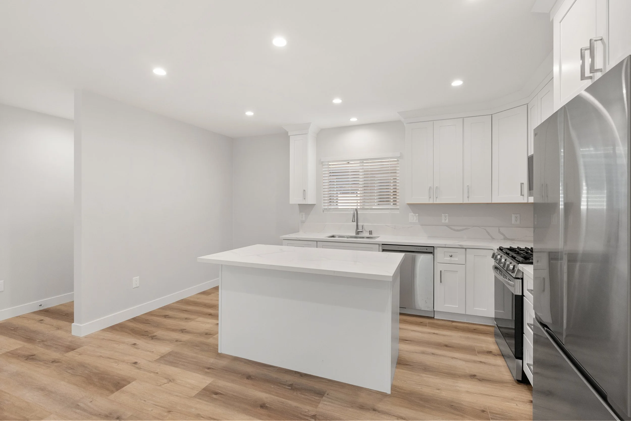 Modern white kitchen with island, hardwood floors, stainless steel appliances, and a window with blinds.