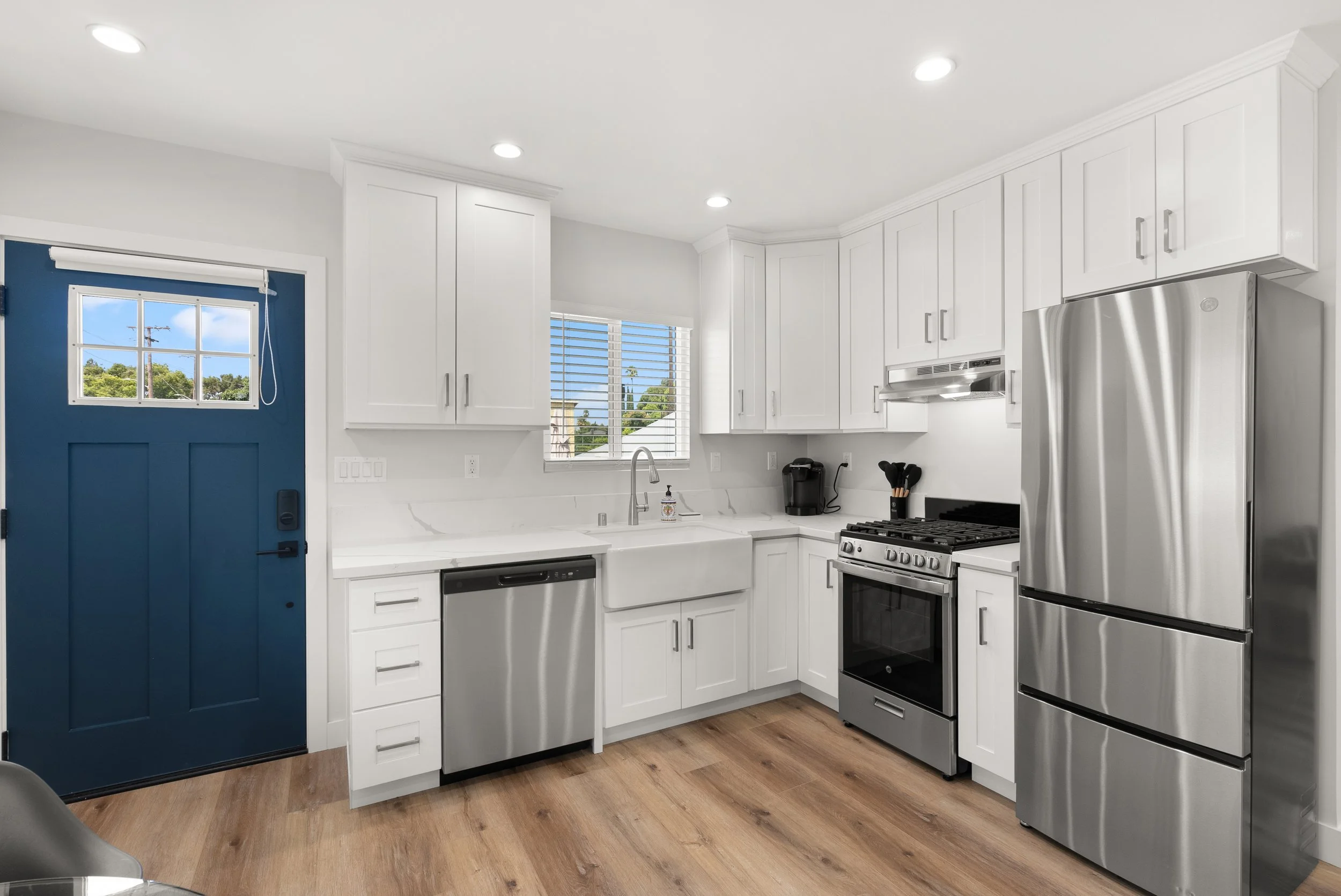 Modern kitchen with white cabinets, stainless steel refrigerator, dishwasher, oven, black coffee maker, and wooden floor, with a blue front door and two windows.