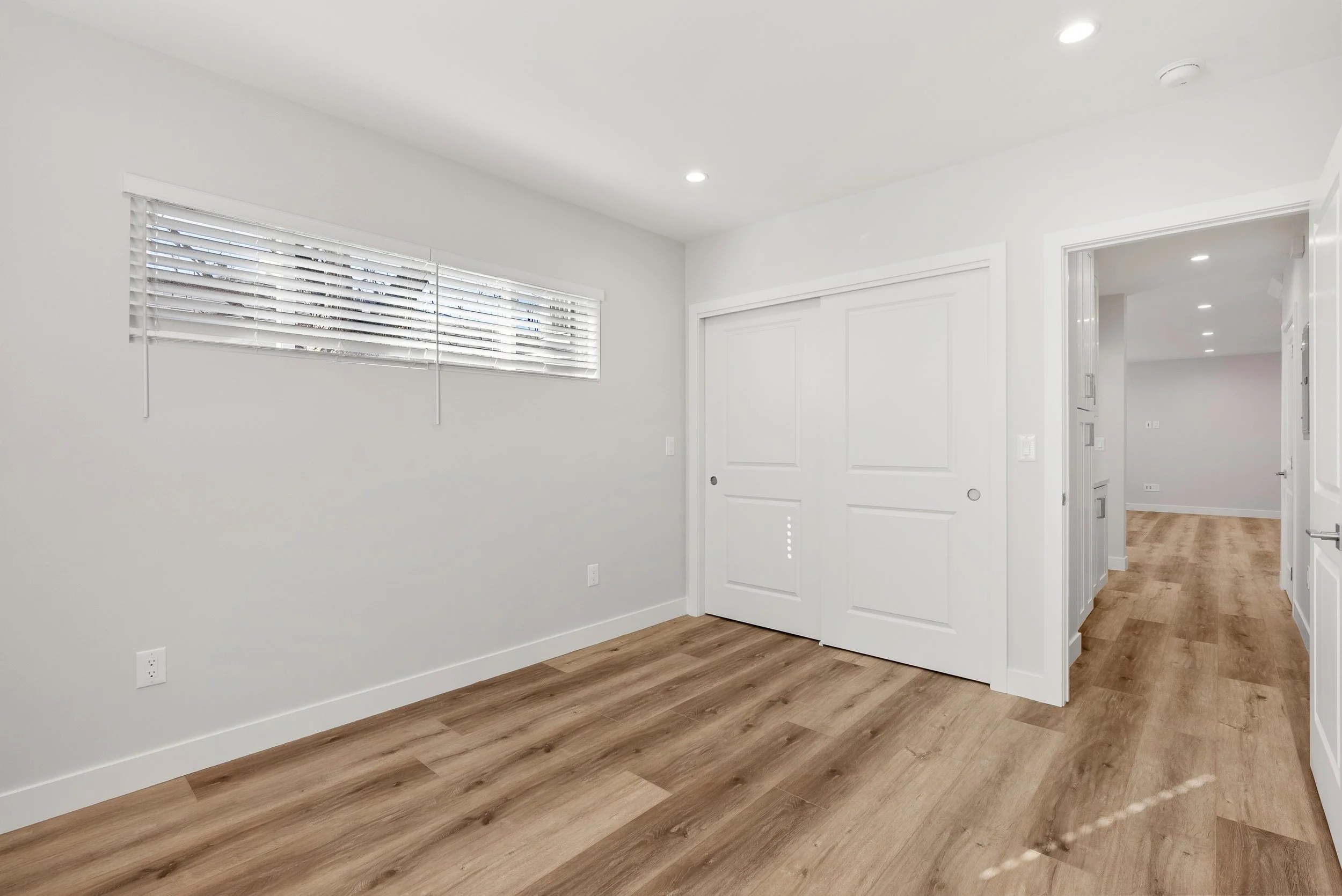 Empty room with wooden floors, white walls, a window with blinds, a white sliding door closet, and an open doorway leading to another room.
