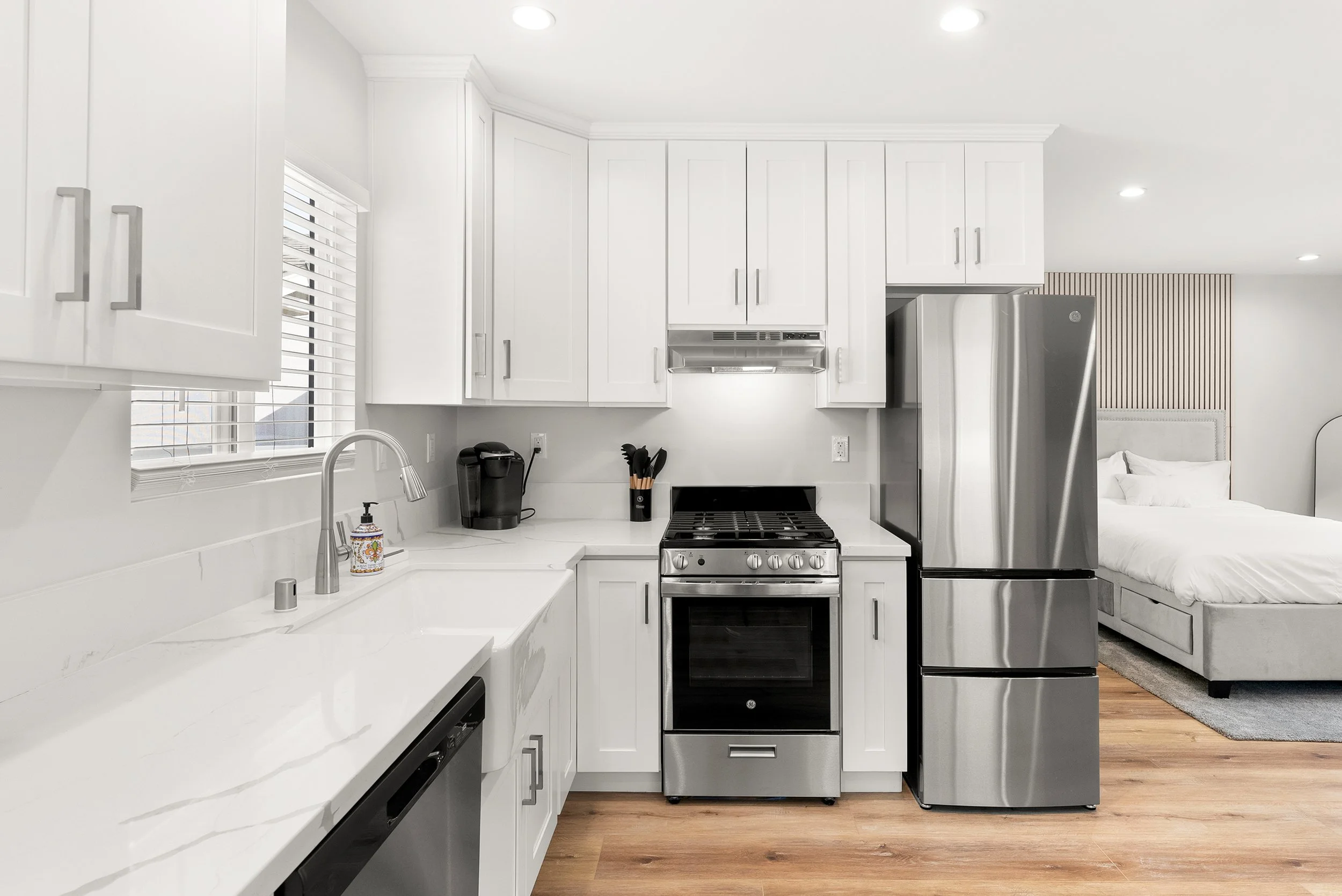 Modern kitchen with white cabinets, stainless steel appliances, and wooden flooring, adjacent to a bedroom with a bed and nightstand.