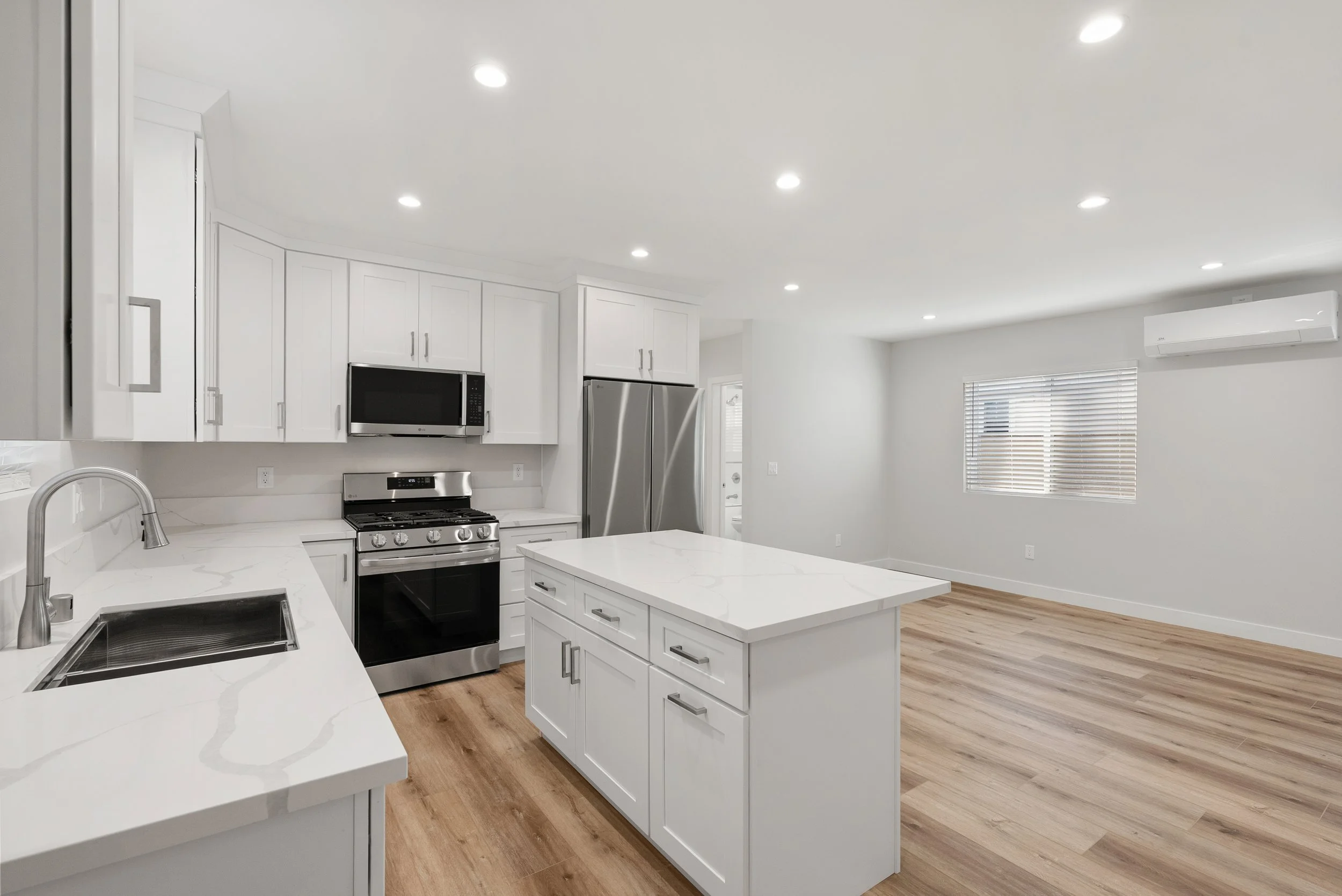 Modern kitchen with white cabinets, stainless steel appliances, island countertop, wood flooring, and a window with blinds.