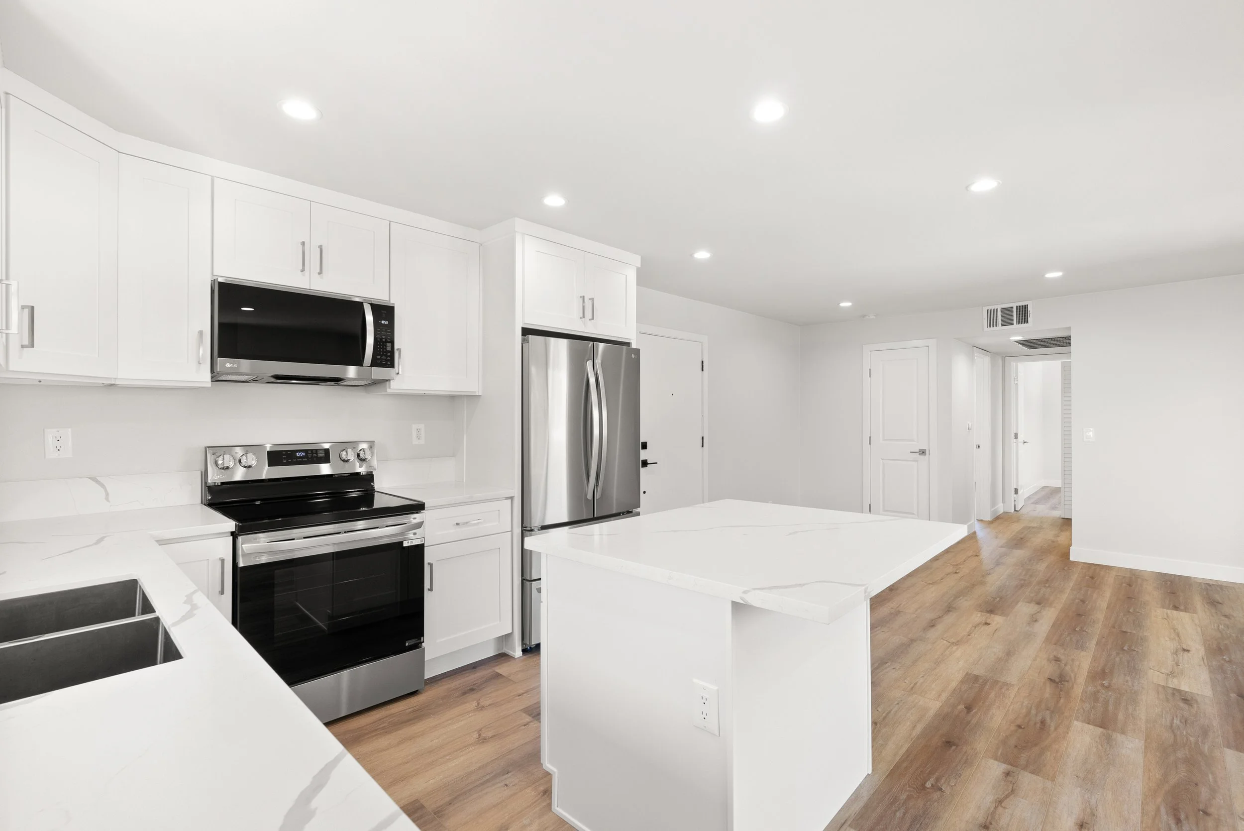 Modern kitchen with white cabinets, stainless steel appliances including a refrigerator and stove, a microwave, and white marble countertops, with wooden flooring and recessed ceiling lights.