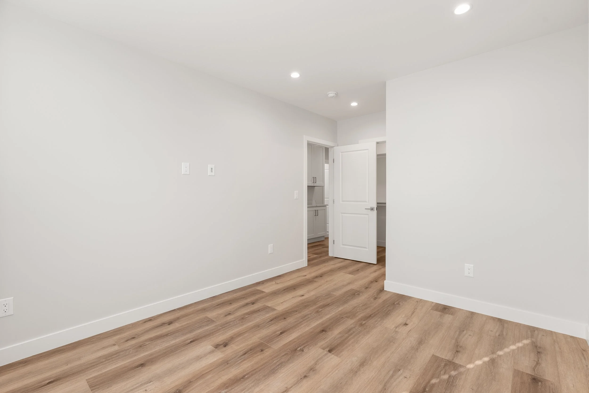Empty room with white walls, wood flooring, and an open door leading to a room with cabinets.