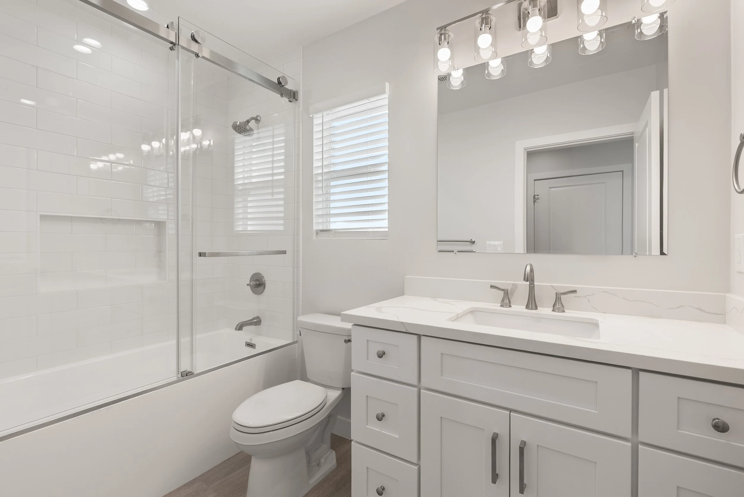 A clean, modern bathroom with a white color scheme, featuring a bathtub with a glass sliding door and a showerhead, a white toilet, a white vanity with a marble countertop, a faucet, drawers, and a cabinet, a large mirror, and a window with white bli