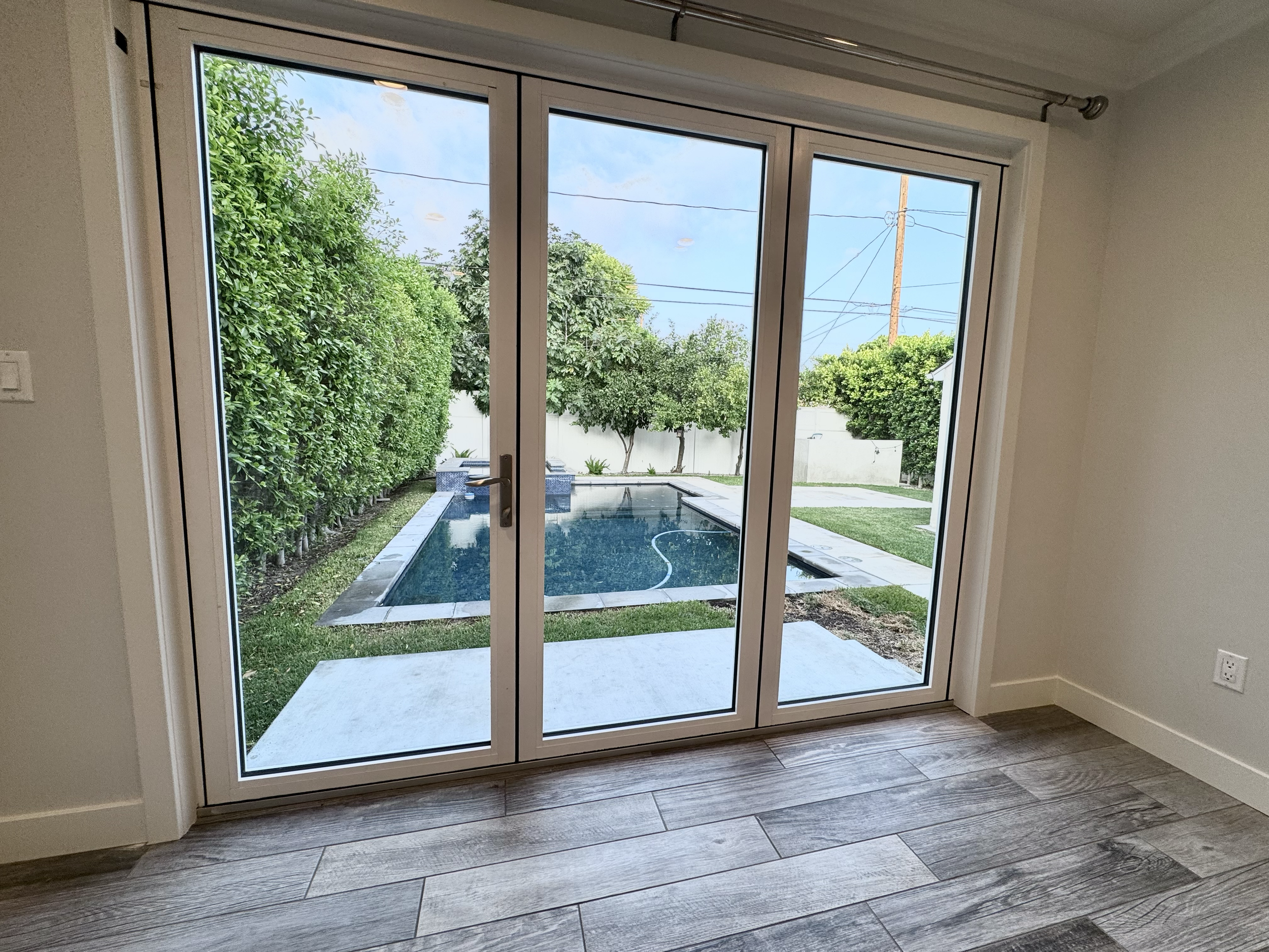 View of a backyard pool through glass sliding doors from inside a room with wooden flooring.