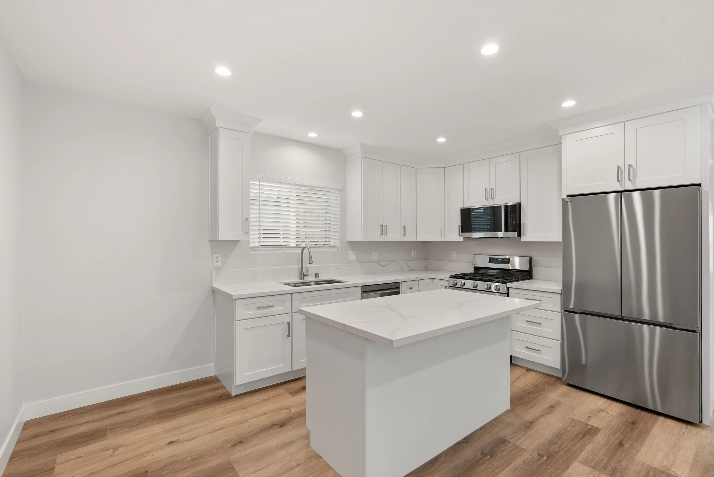 Modern kitchen with white cabinets, marble countertops, stainless steel appliances including a refrigerator and oven, and hardwood flooring.