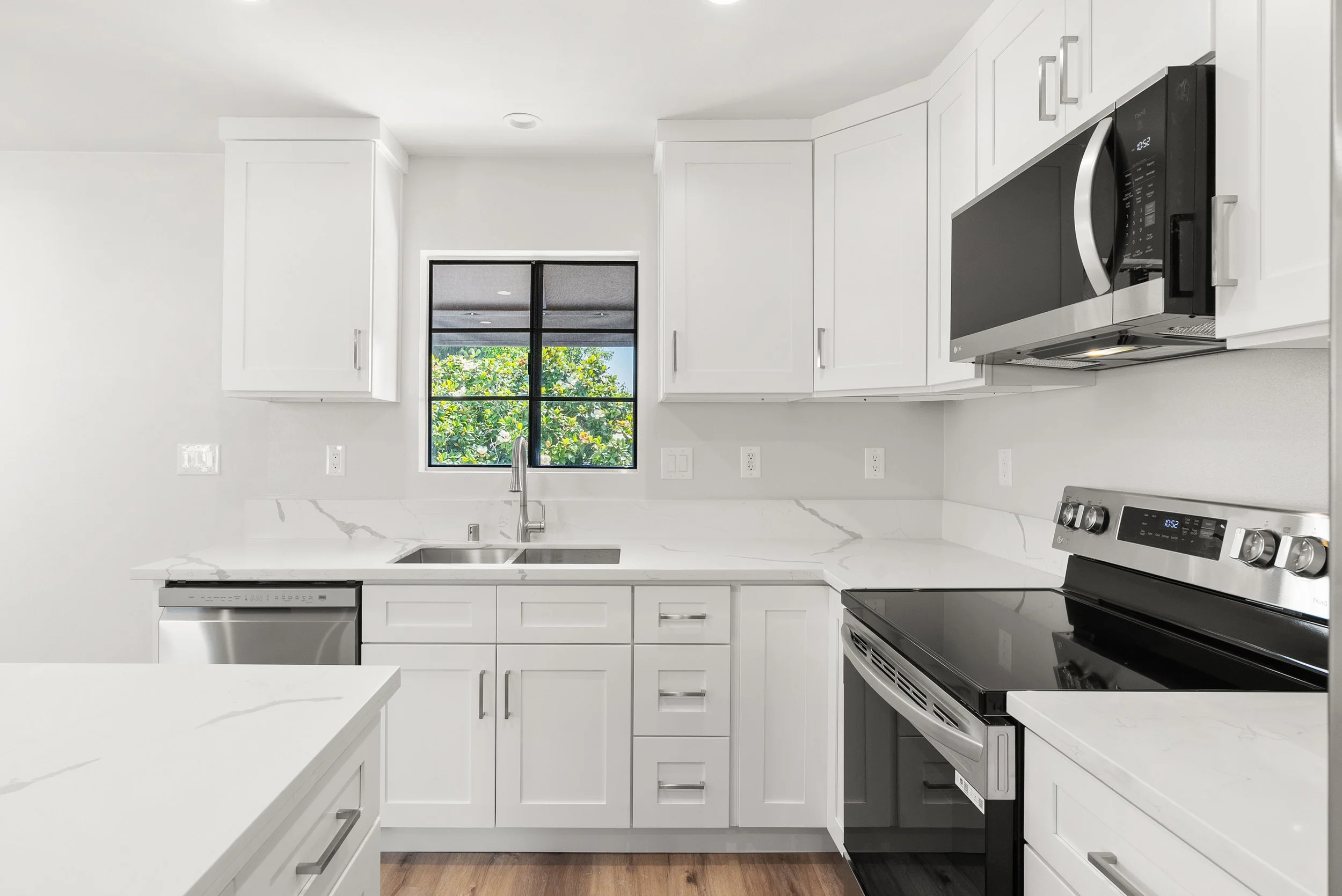 Modern white kitchen with marble countertops, stainless steel appliances, and a window overlooking greenery.