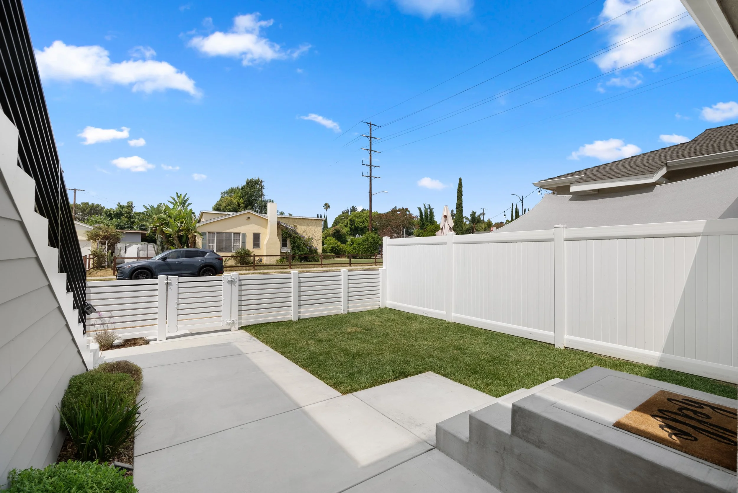 View of a backyard with white privacy fence, green lawn, concrete patio, and steps with a welcome mat, under a blue sky with clouds.