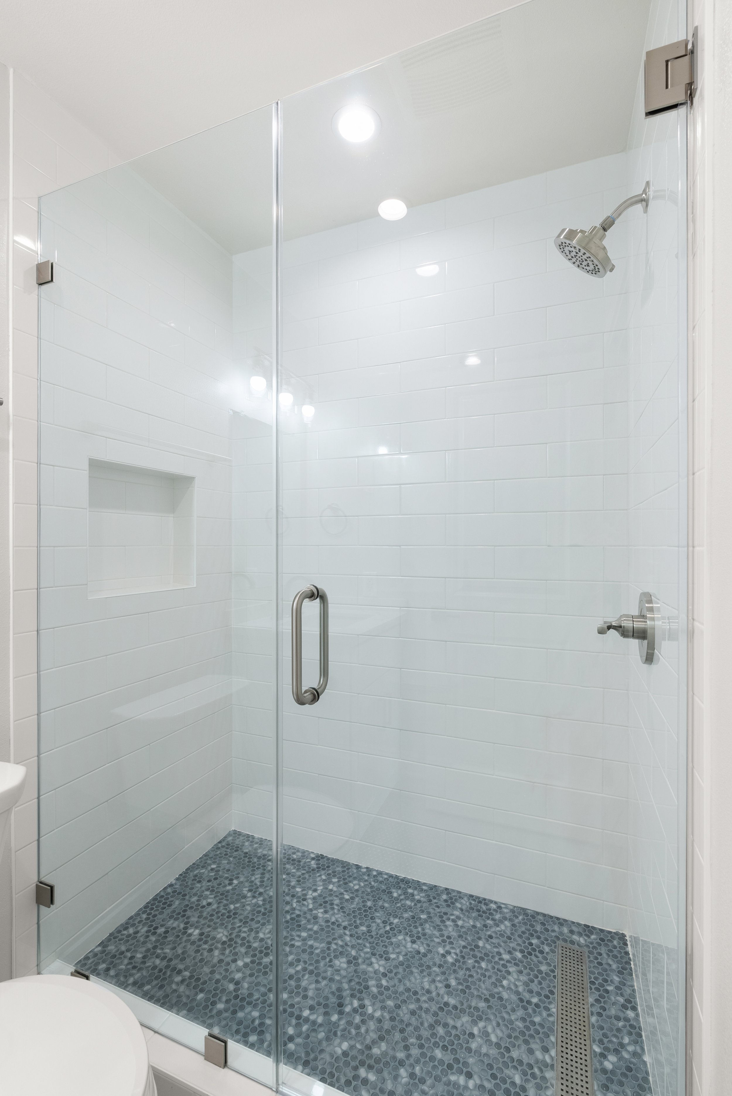 Clean glass shower enclosure with white tiled walls, a built-in niche, and pebble tile floor, with a showerhead and controls.