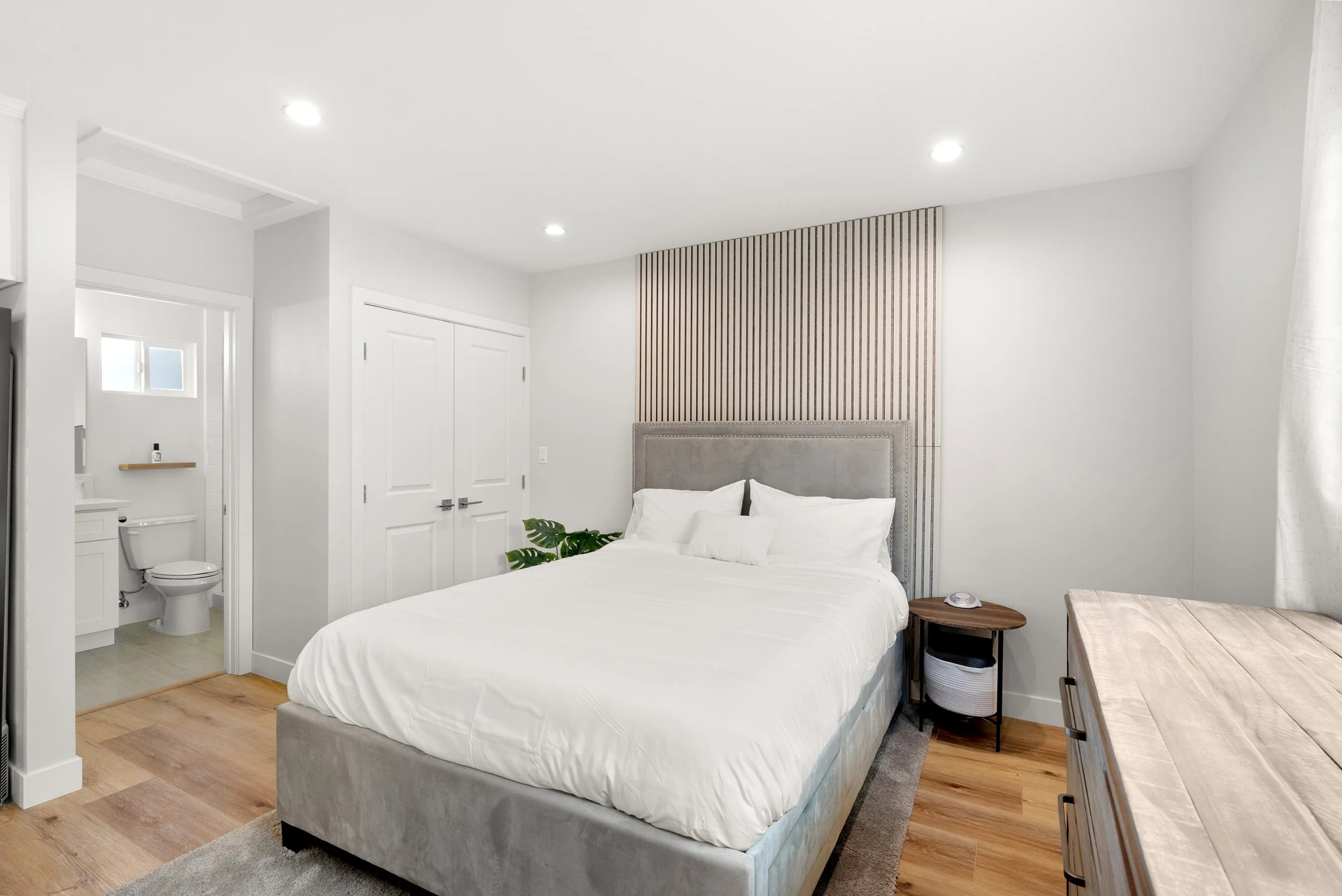 Bedroom with a bed featuring a grey upholstered headboard, white bedding, and pillows. A small round side table with a plant next to the bed, and a wooden dresser on the right. A bathroom with a toilet and small window visible through an open door on