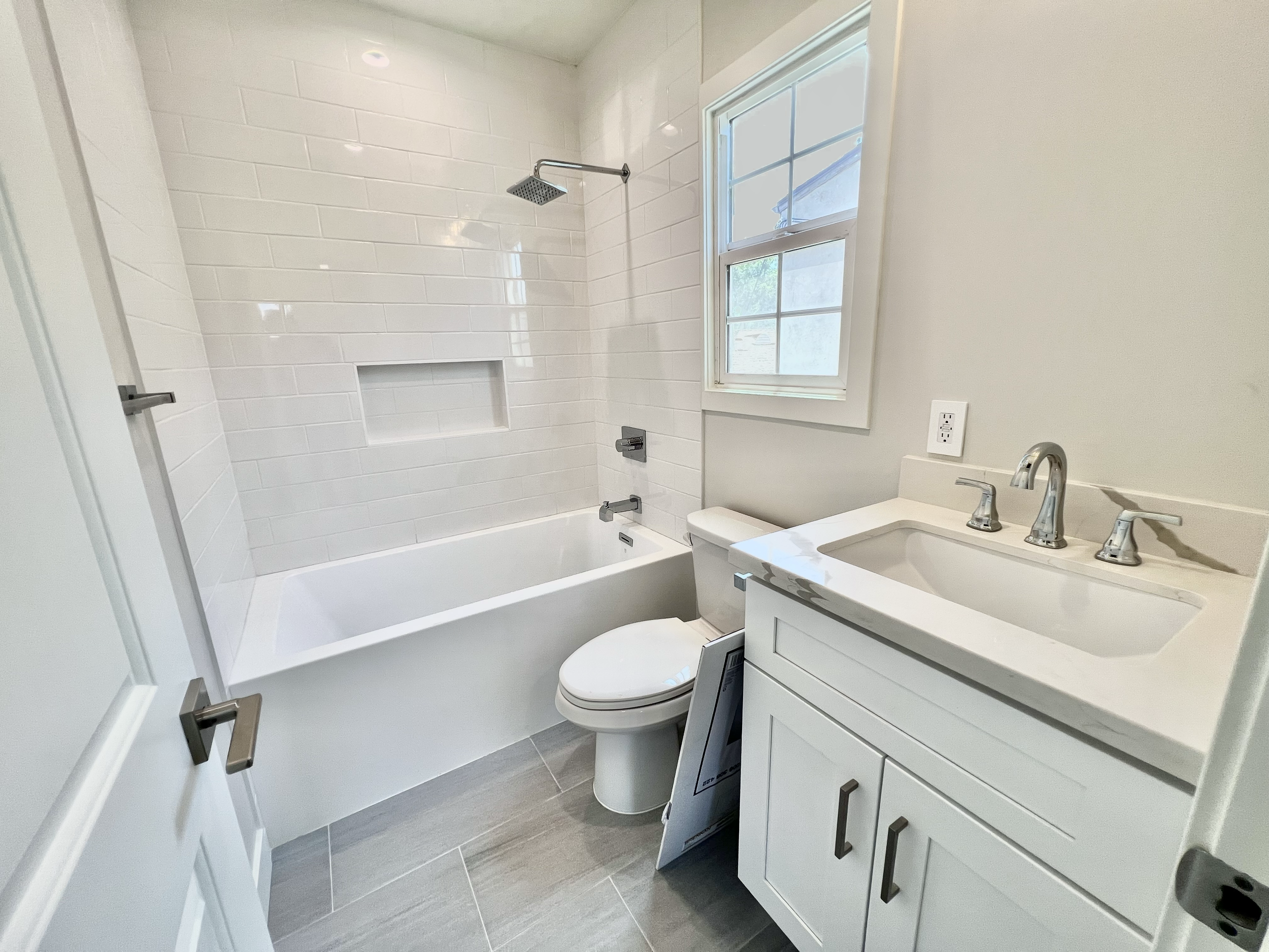 Modern bathroom with white tile bathtub and shower, white toilet, and white vanity with sink. There is a window with divided panes above the toilet, and the floor has gray tiles.