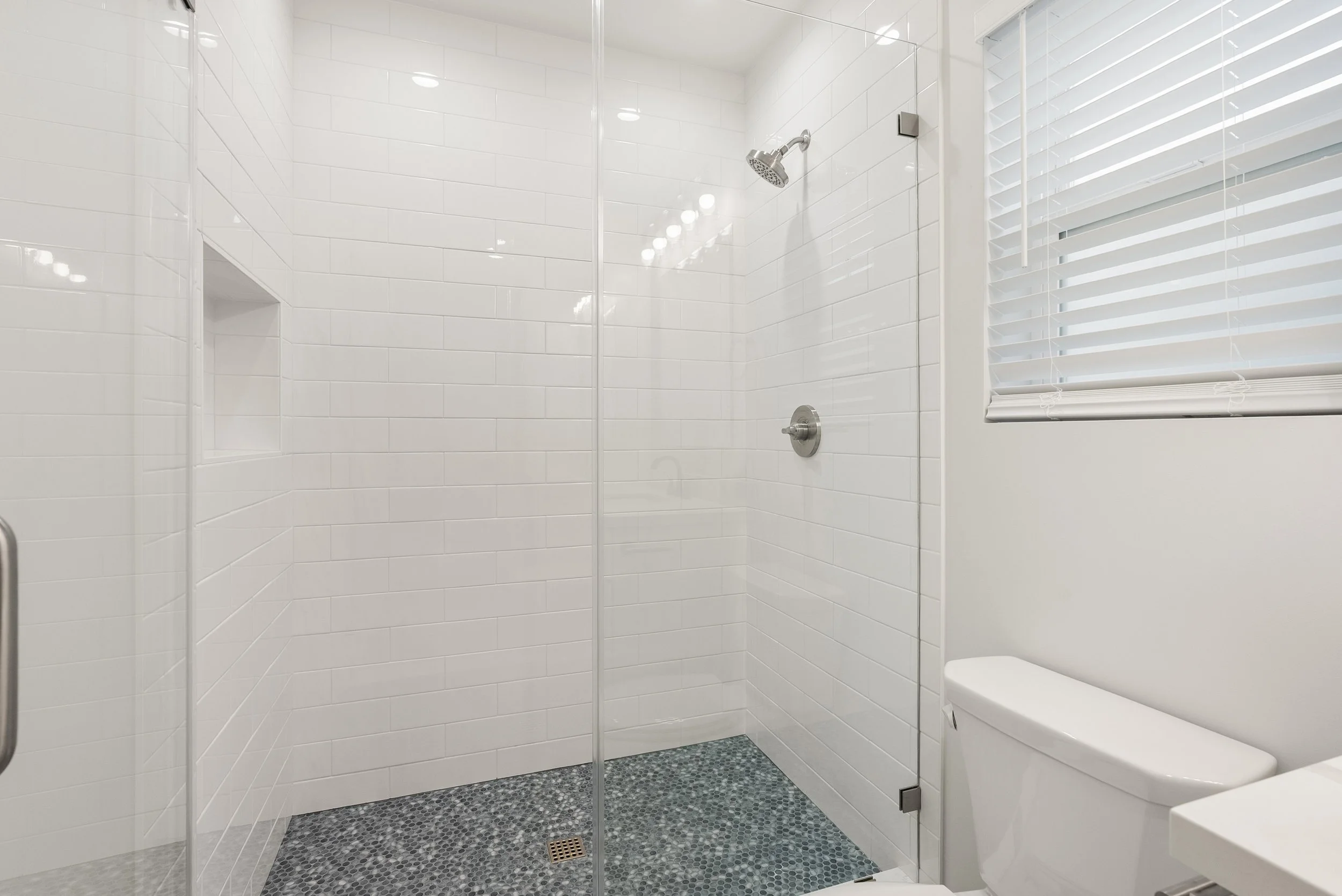 Modern bathroom shower with white subway tiles, glass enclosure, small pebble floor, and a window with white blinds.