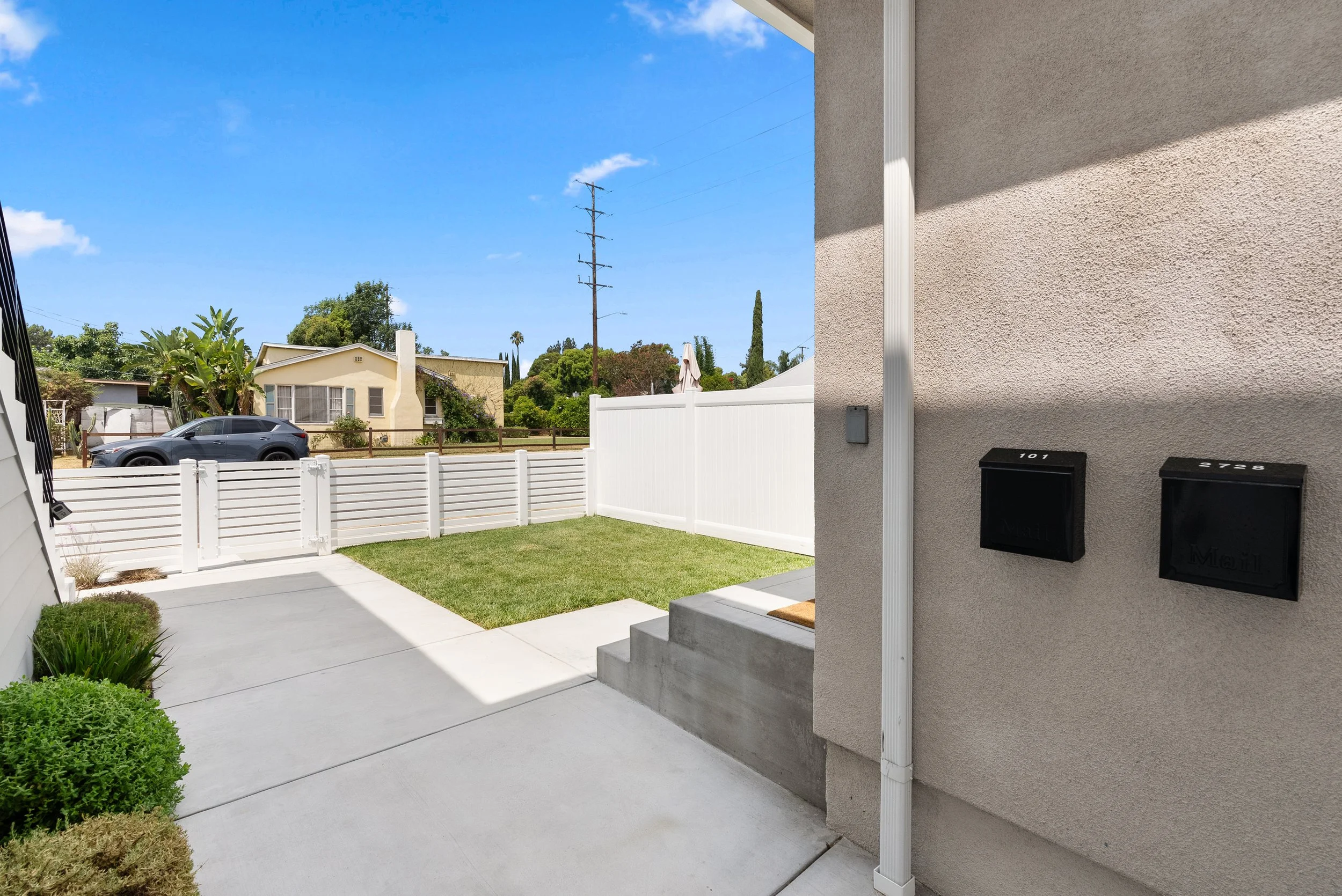 View of a front yard with a concrete sidewalk and stairs, a small grassy lawn, a white fence, a parked gray car on the street, neighboring houses, a utility pole, and a clear blue sky.