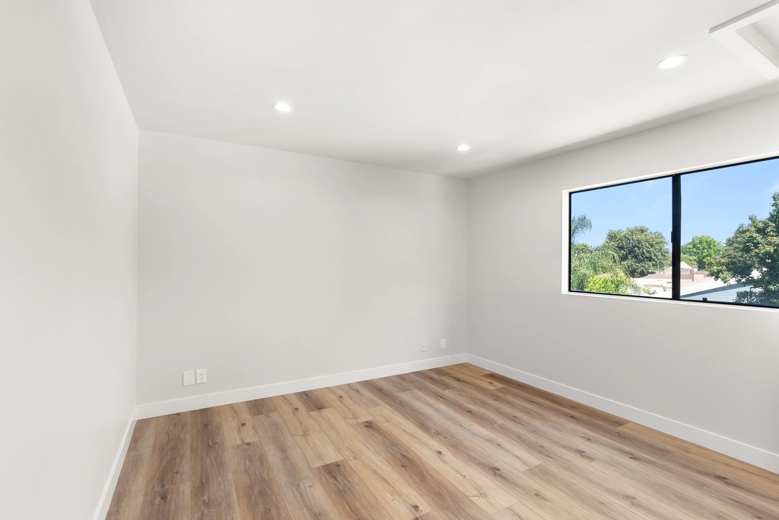 Empty room with white walls, hardwood flooring, a large window showing trees and a blue sky, and recessed ceiling lights.