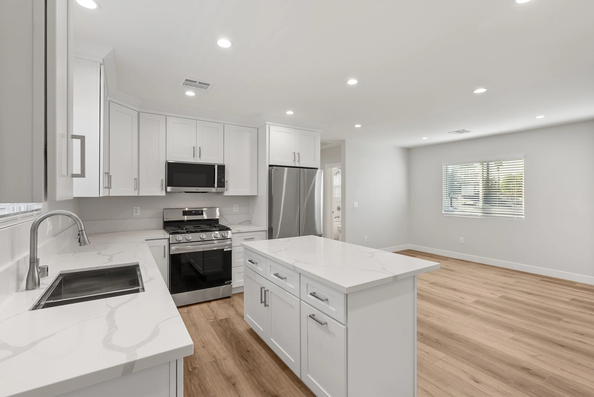 Modern kitchen with white cabinets, marble countertops, stainless steel appliances, and light wood flooring, with an open layout and window with blinds.