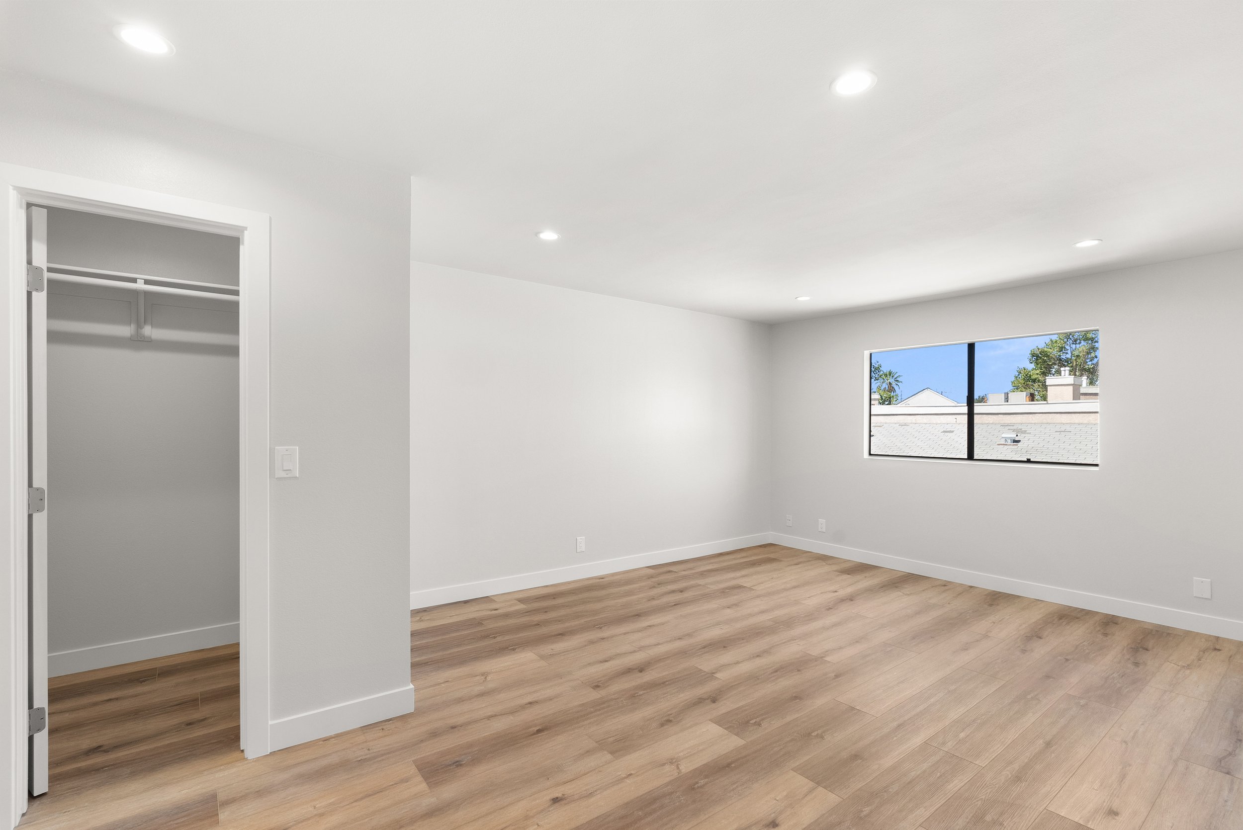 Empty room with hardwood flooring, white walls, a window showing a view of rooftops and trees, and an open closet