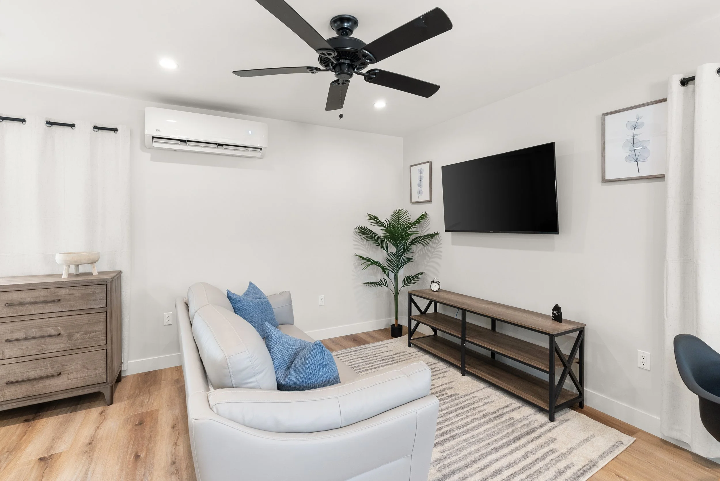 Living room with white walls, wooden flooring, a white sofa with blue cushions, a wooden TV stand, mounted flat-screen TV, potted plant, ceiling fan, air conditioner, framed art, and beige curtains.