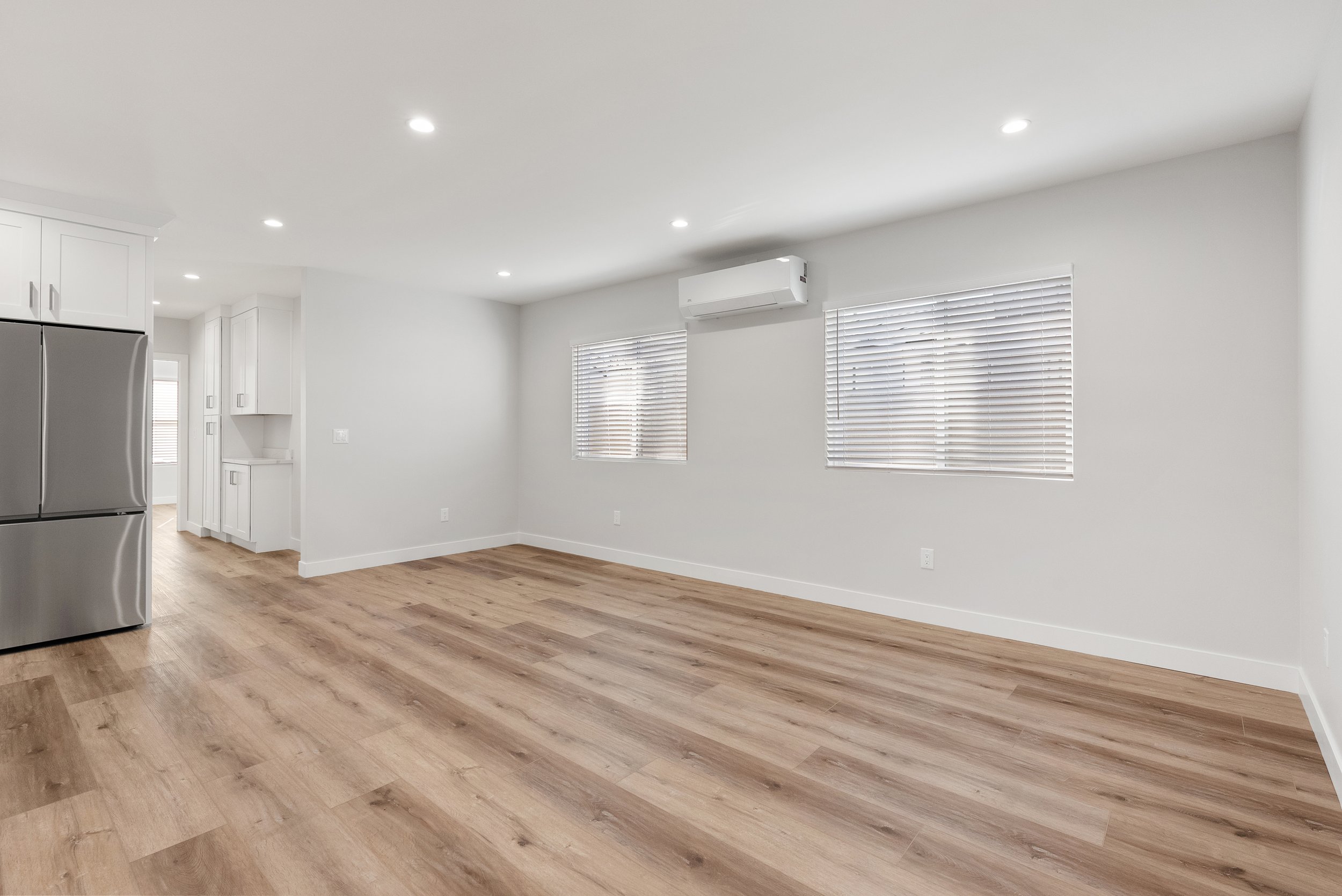 Empty living room with white walls, wooden flooring, two windows with blinds, and a wall-mounted air conditioning unit. Part of a kitchen with a stainless steel refrigerator is visible on the left.