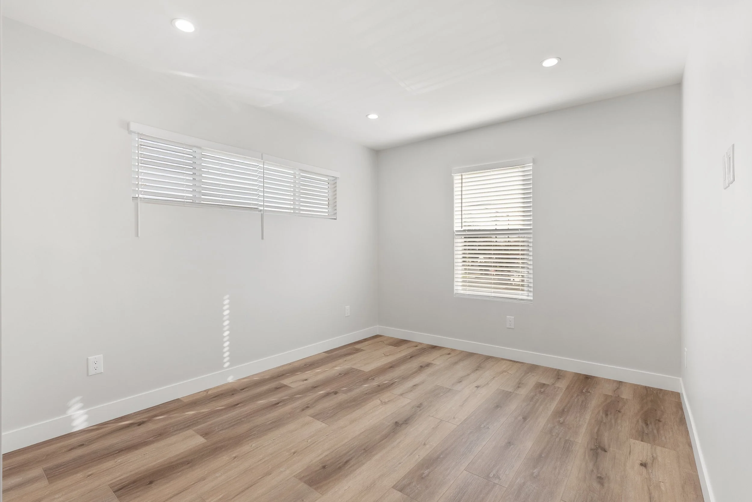 Empty room with white walls, two windows with blinds, wooden floor, and ceiling lights.