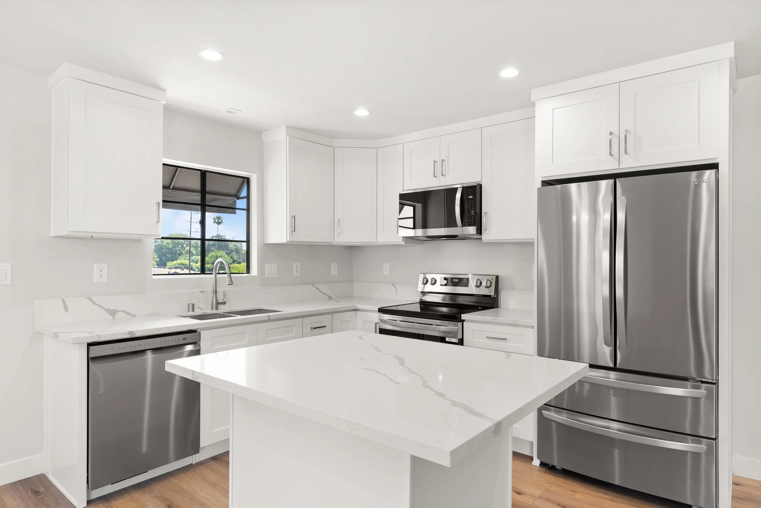 Modern white kitchen with stainless steel appliances, marble countertops, and a window view of greenery outside.