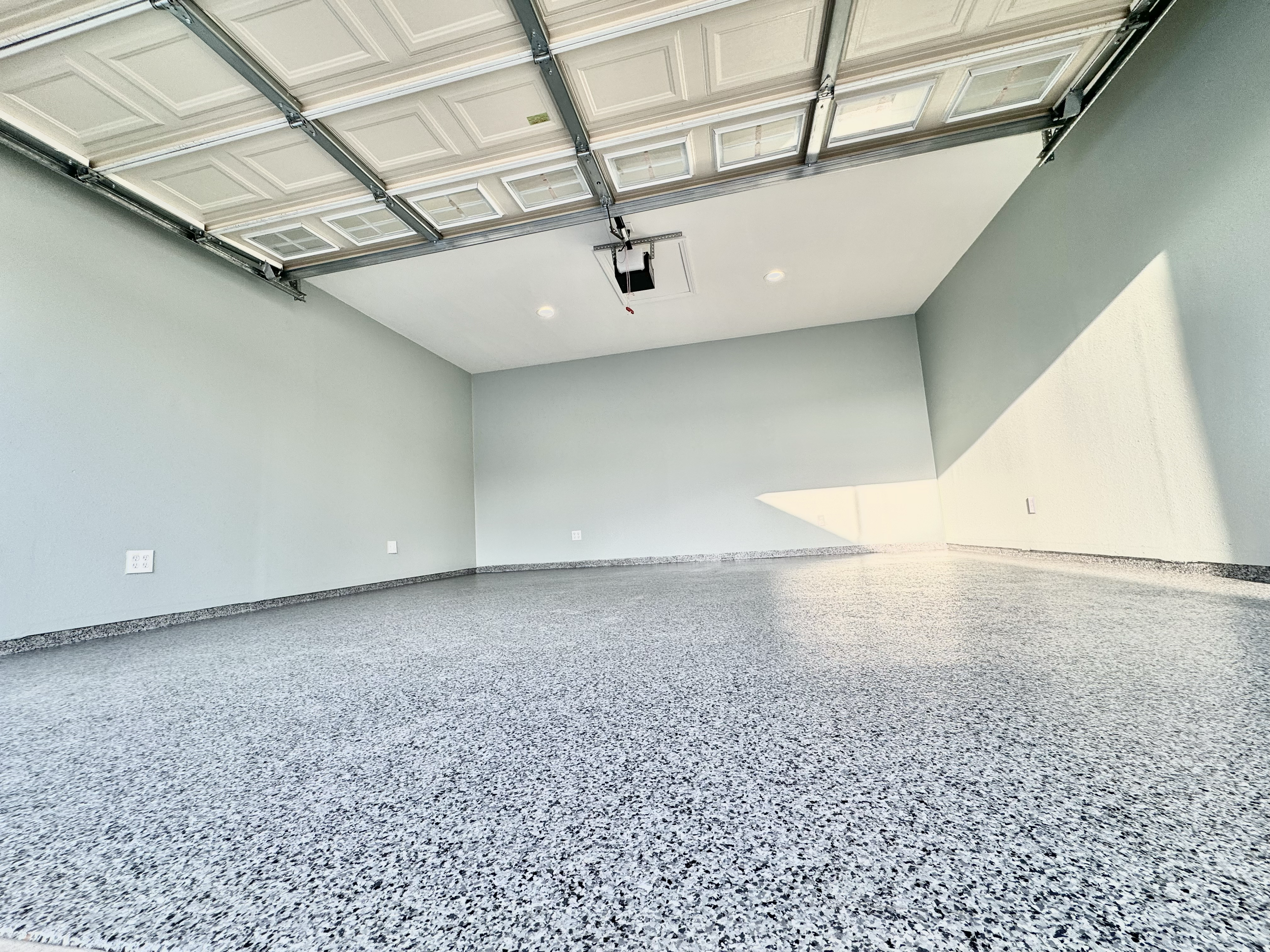 Empty garage with a speckled floor, light green walls, a partially opened garage door, and ceiling lights.